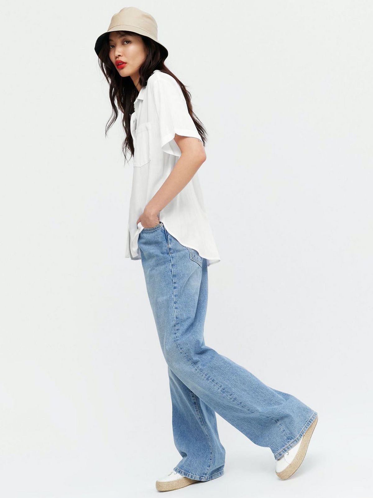 Woman wearing a bucket hat, off shite button up shirt, blue jeans and trainers