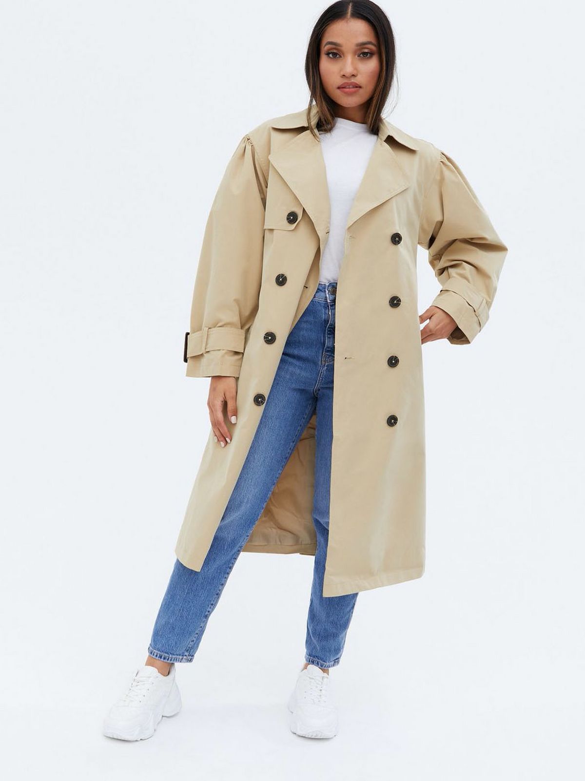 Woman wearing a stone puff sleeve belted trench coat, white top, jeans and trainers