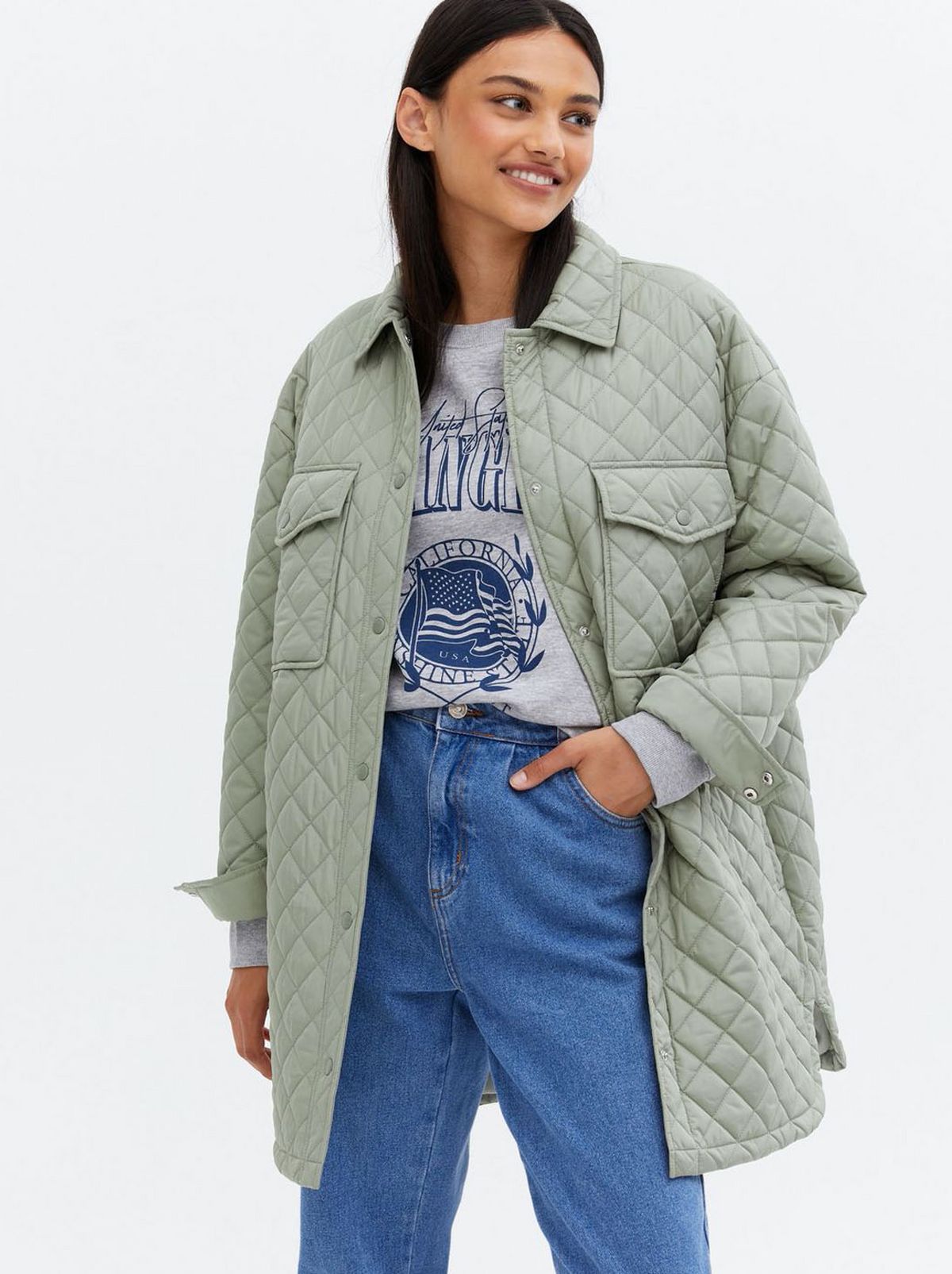 Woman wearing a light green quilted front shacket, grey logo t-shirt and blue jeans