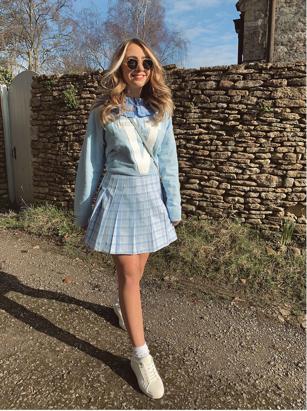 woman walks on street, wears light blue varsity sweatshirt, light blue tennis skirt and white trainers