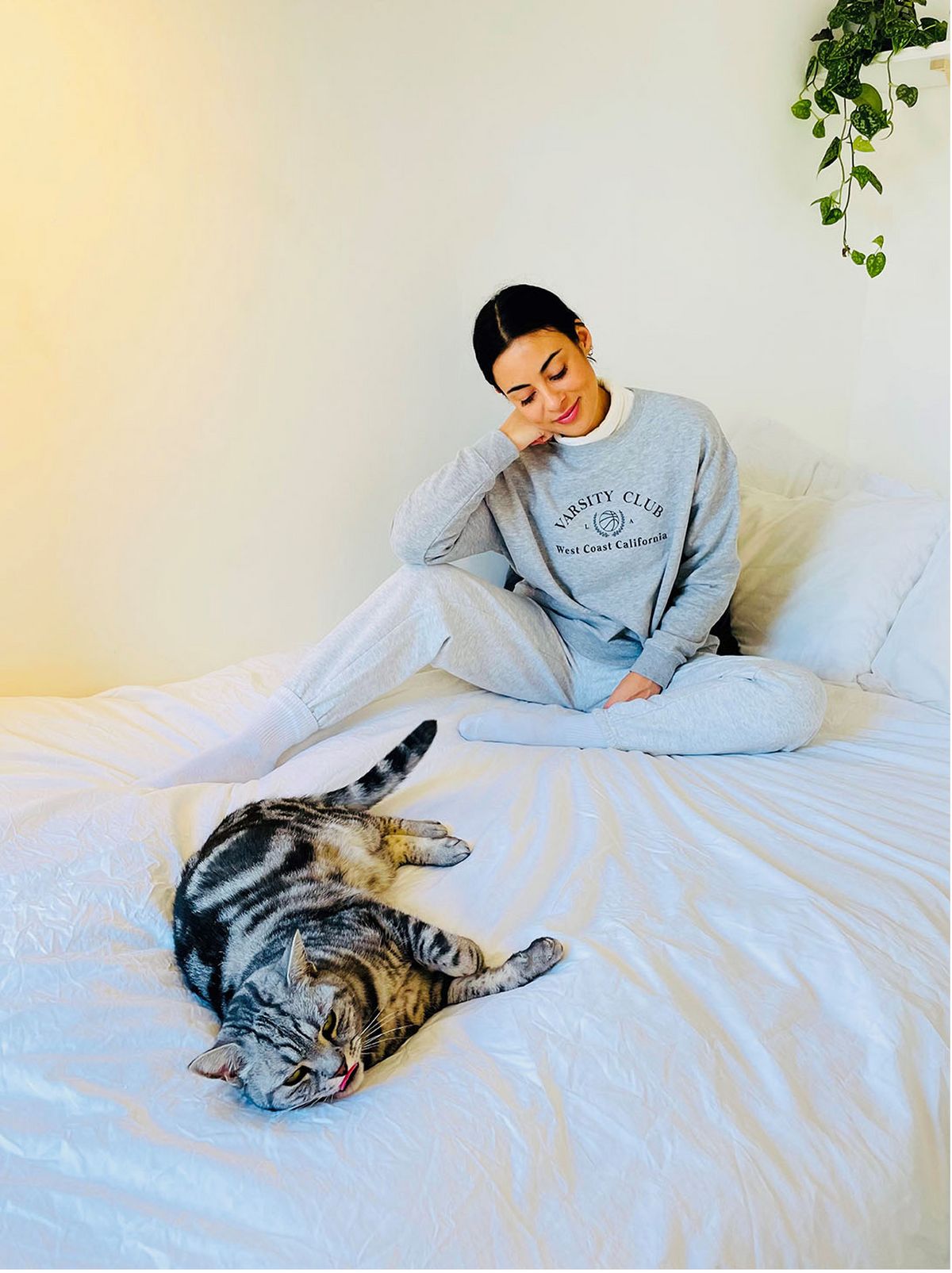 woman sits on bed next to grey cat, wears varsity club logo grey sweatshirt and grey joggers