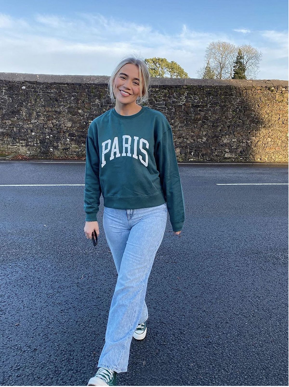 woman crosses road, wears pine green sweatshirt with paris logo, blue jeans and green plimsolls