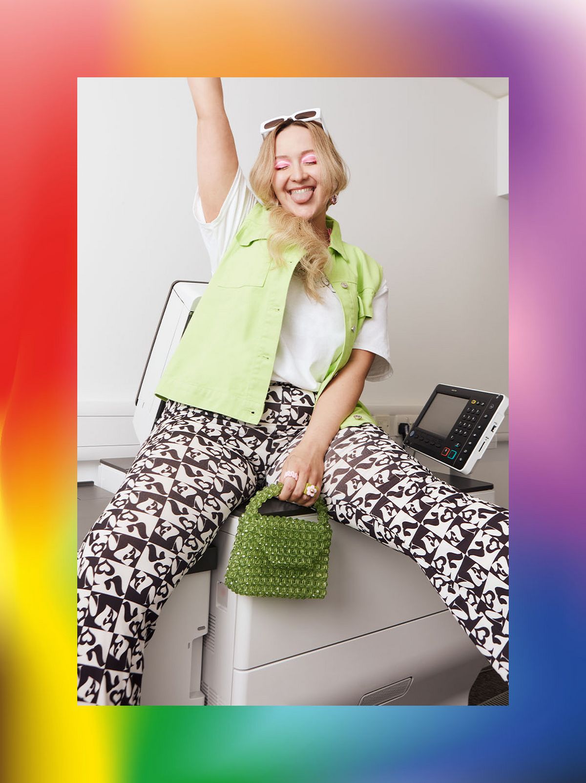 Lucinda Poulson sits on a printer smiling with her arm raised wearing a lime green vest jacket and a pair of black & white printed trousers	