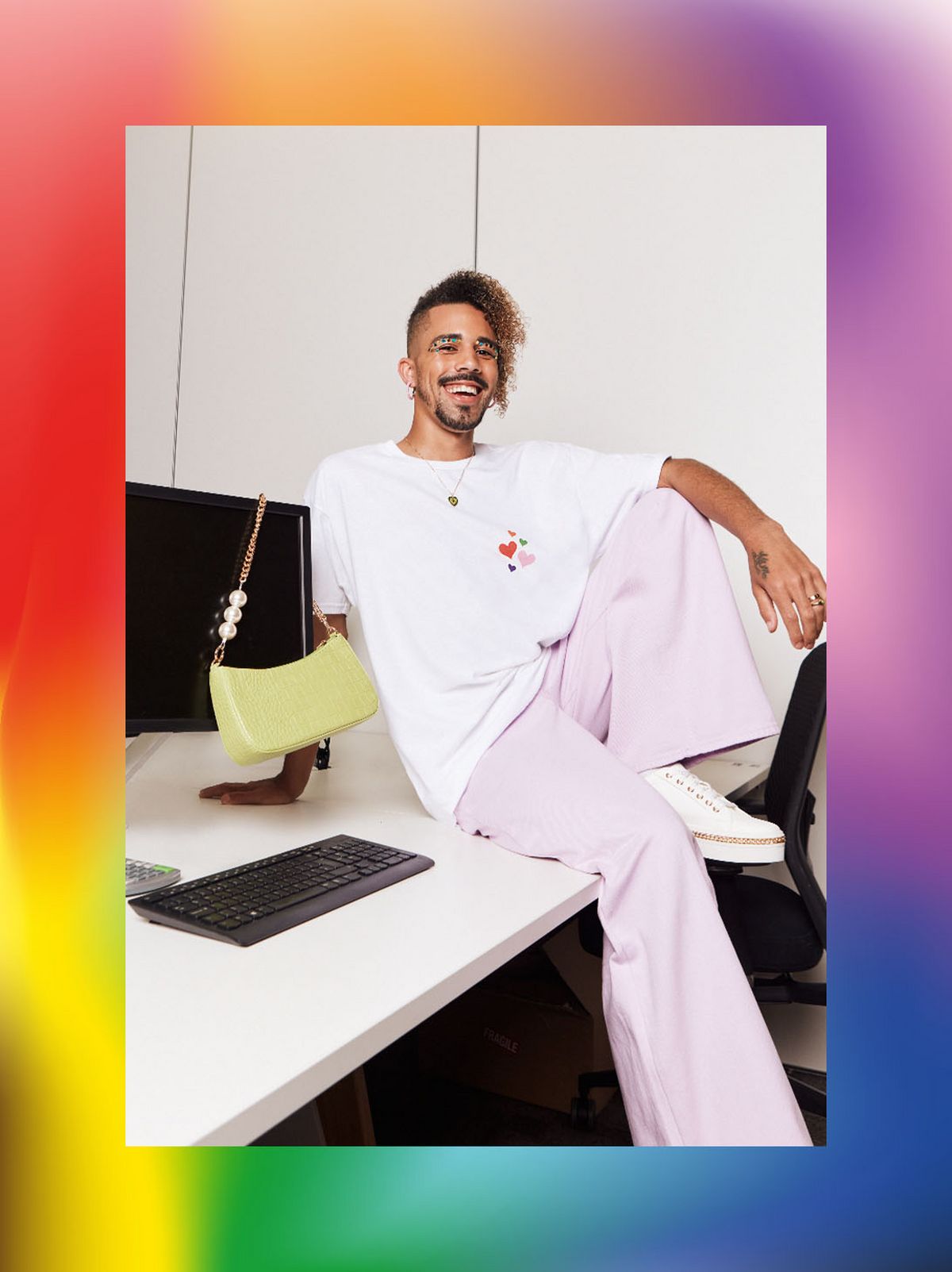 Daniel Junior Harrison is sitting on a desk, laughing and wearing a pair of straight leg light pink trousers and a white T shirt	