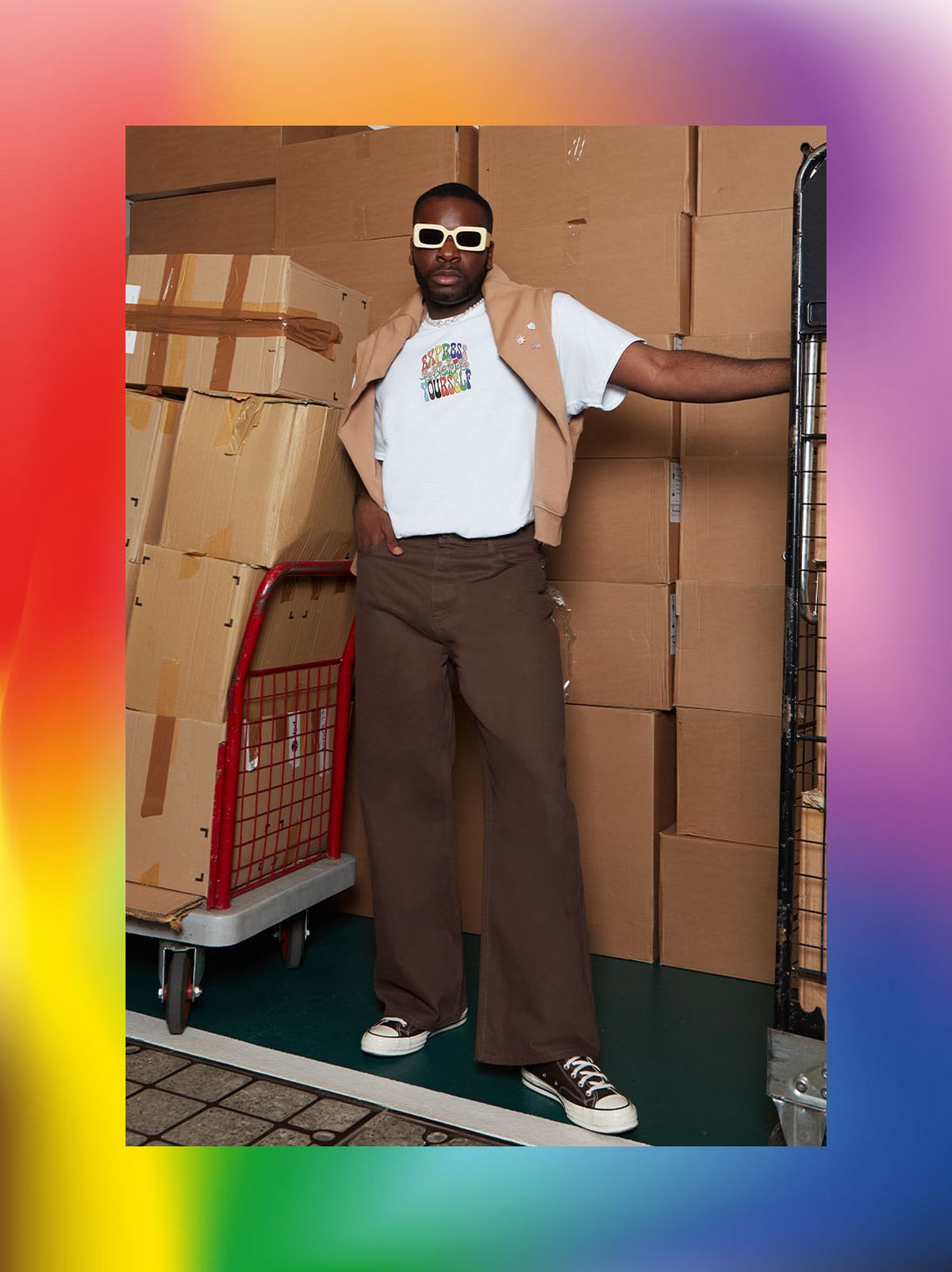 Theo A-Regisford is staring at the camera, wearing a pair of wide fit brown trousers, a white print t shirt and a beige gilet	