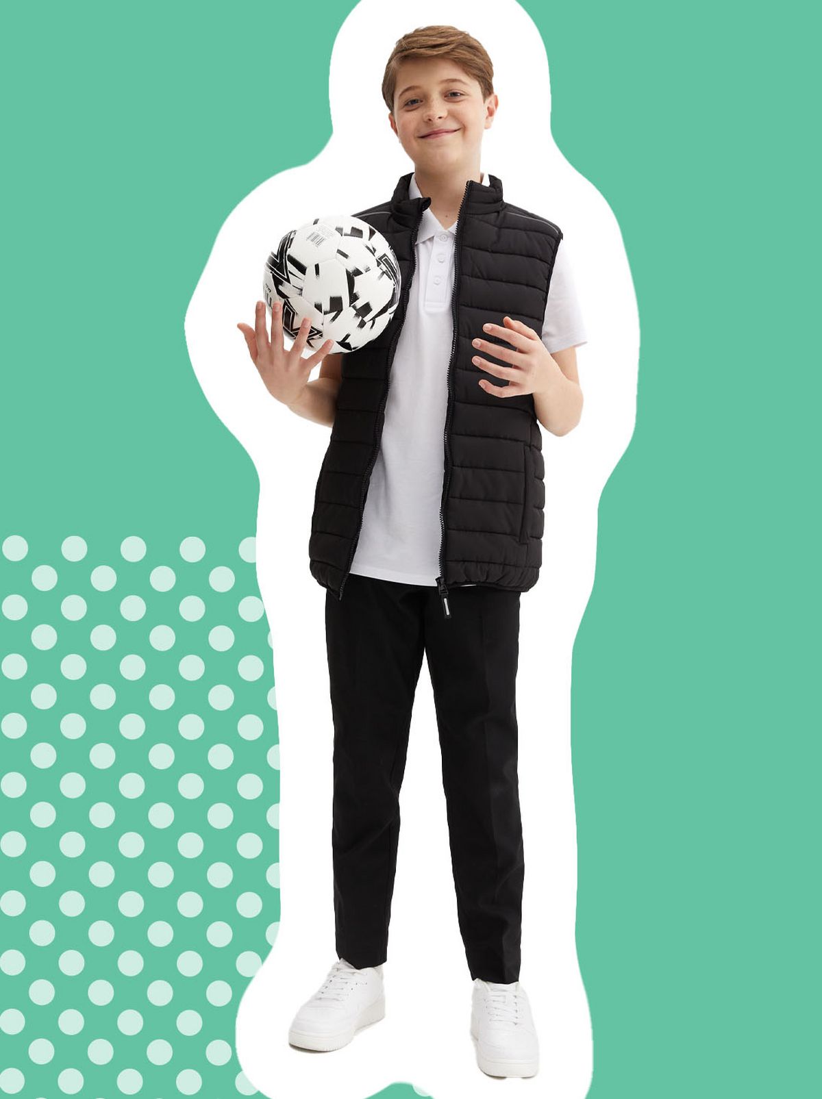 A boy is standing holding a football wearing a light jacket, a pair of school trousers and a school polo shirt