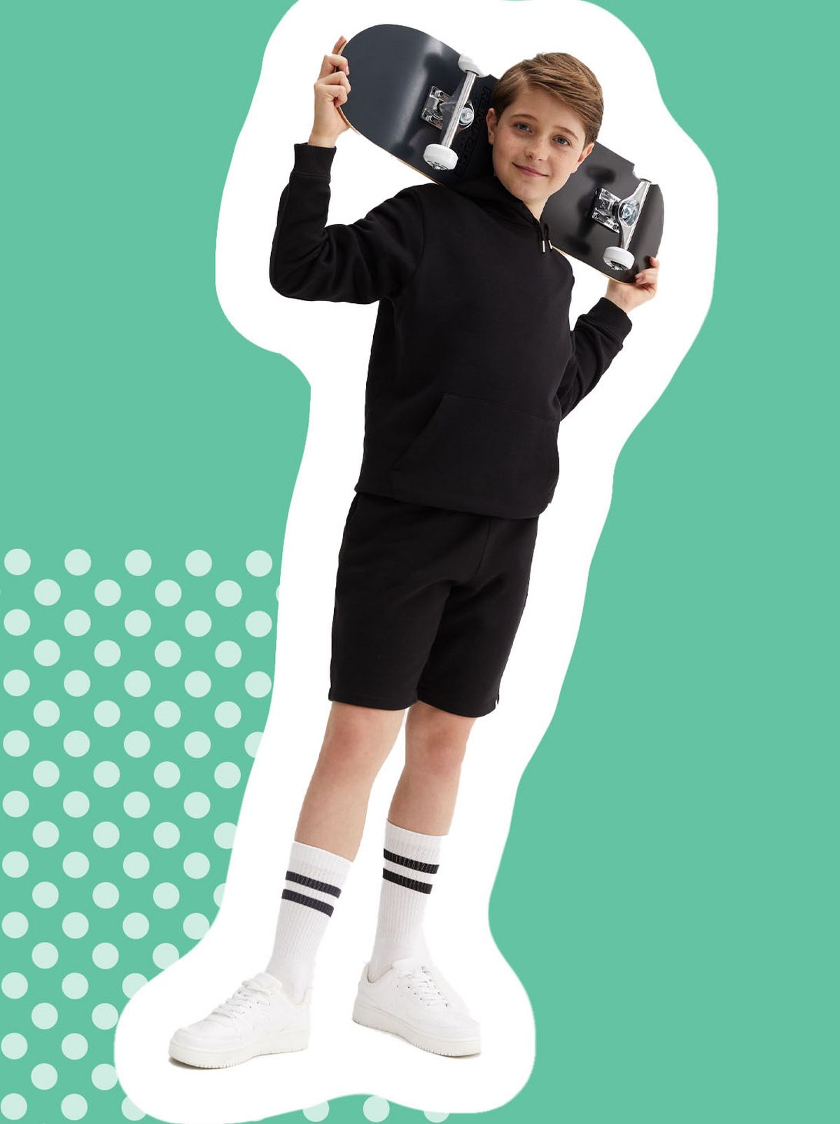 A boy is holding a skateboard, wearing a black jumper and black pe shorts