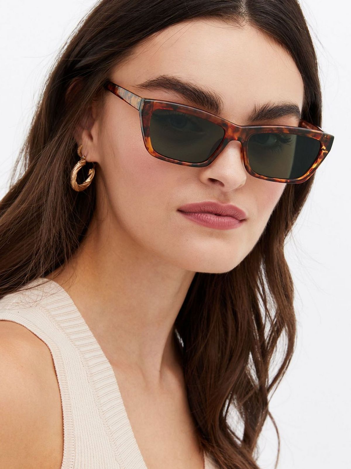 We see a women wearing a pair of sunglasses and a cream vest, staring at the camera in front of a white background