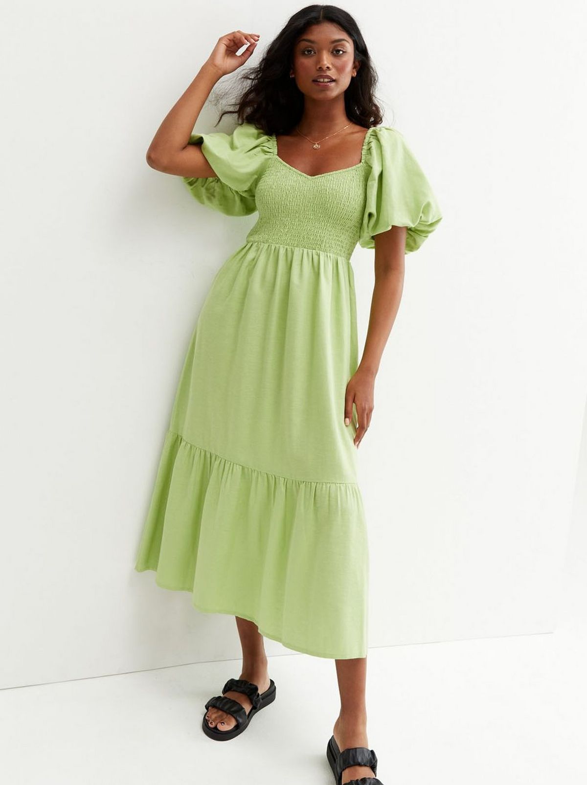 There is a women standing in front of a white backdrop wearing a green and white checked dress and a pair of strappy sandals