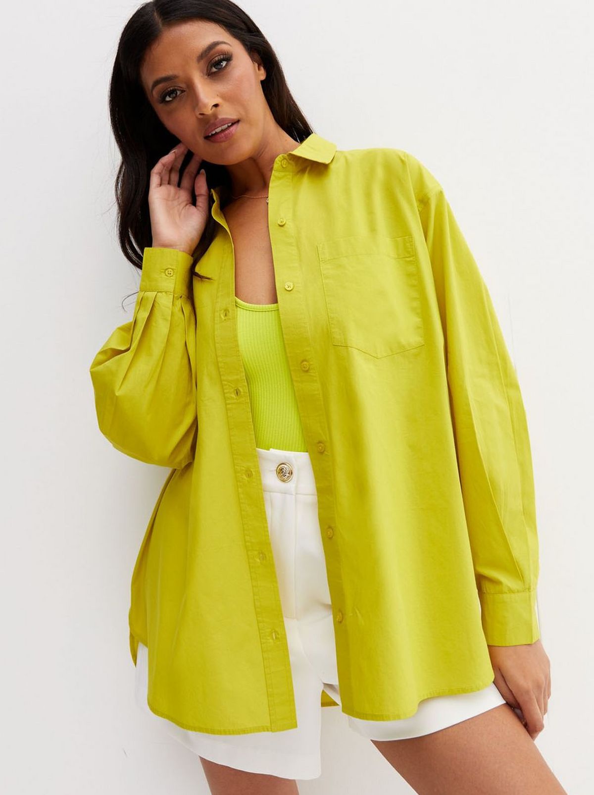 There is a women that wears a long sleeve yellow shirt and a pair of white shorts, looking at the camera touching her ear looking at the camera
