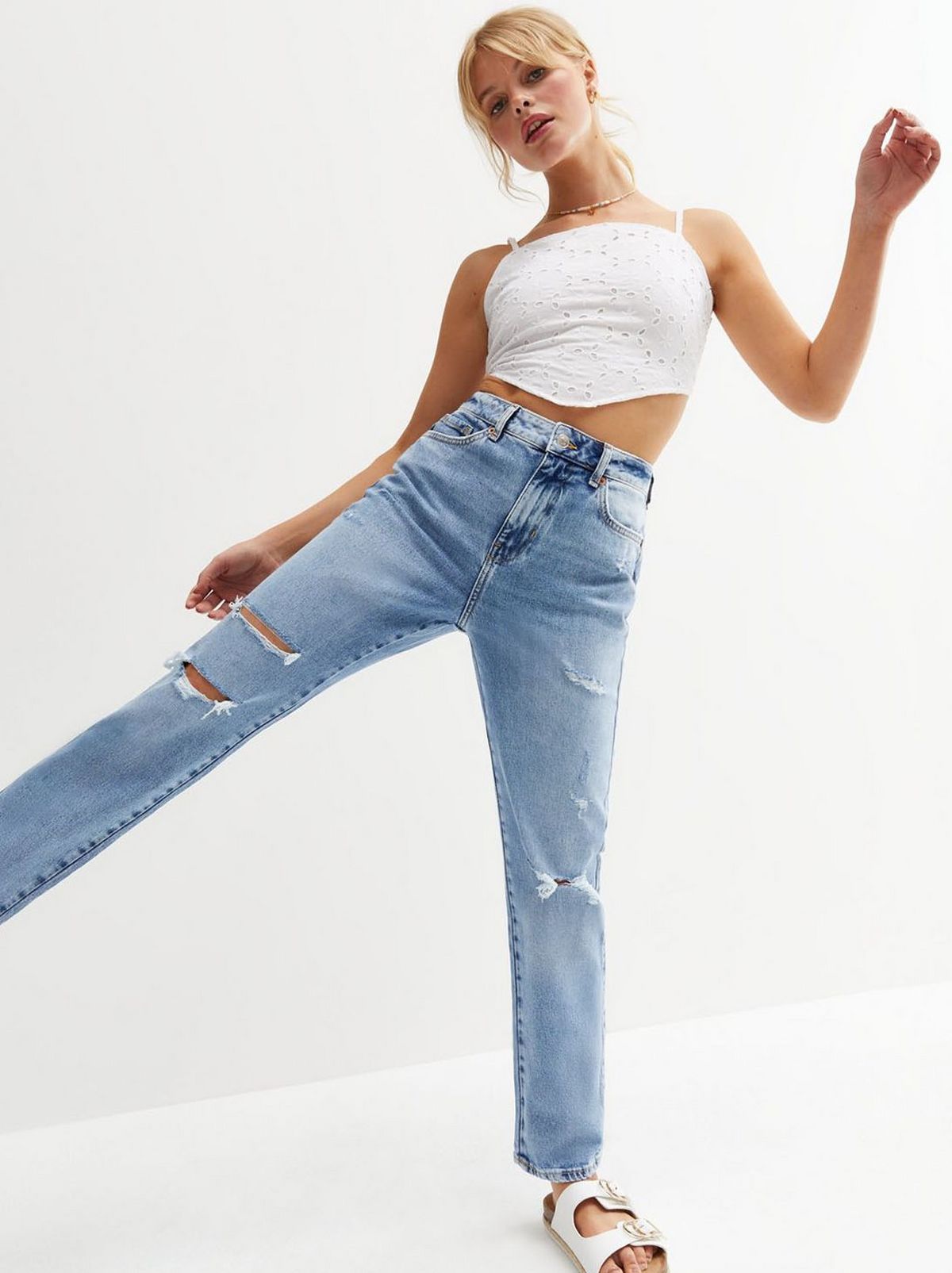There is a women, balancing on one leg with her other leg outstretched to the side, who looks at the camera while she wears a pair of ripped denim jeans and a white vest