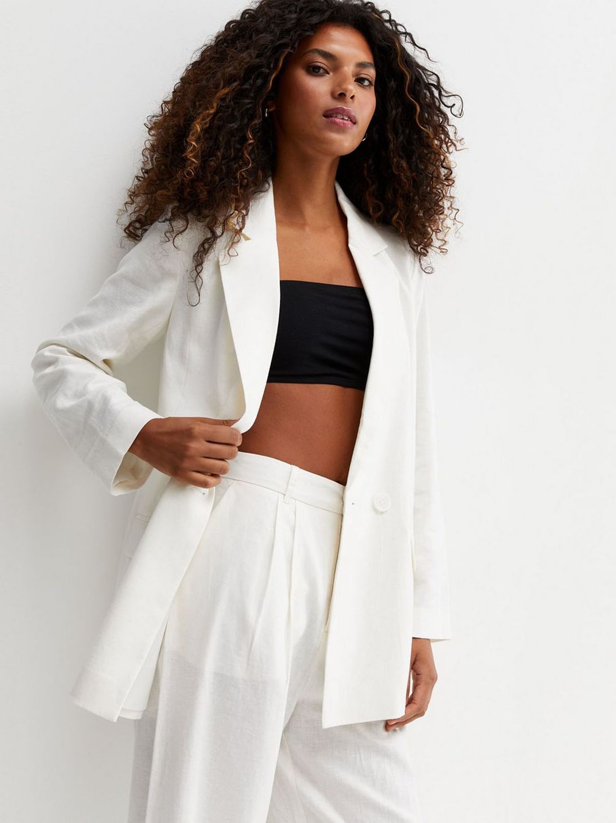 There is a women leaning on a white wall, staring at the camera, wearing a white blazer and loose white trousers and a black band crop top
