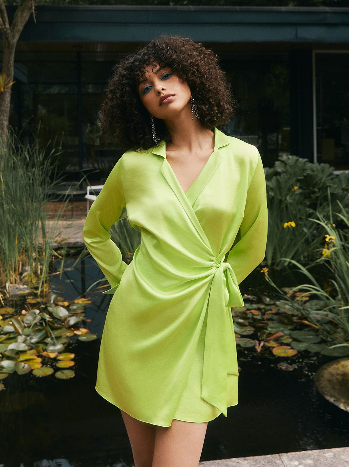 There is a women wearing a lime green mini dress, standing looking at the camera in front of a pond