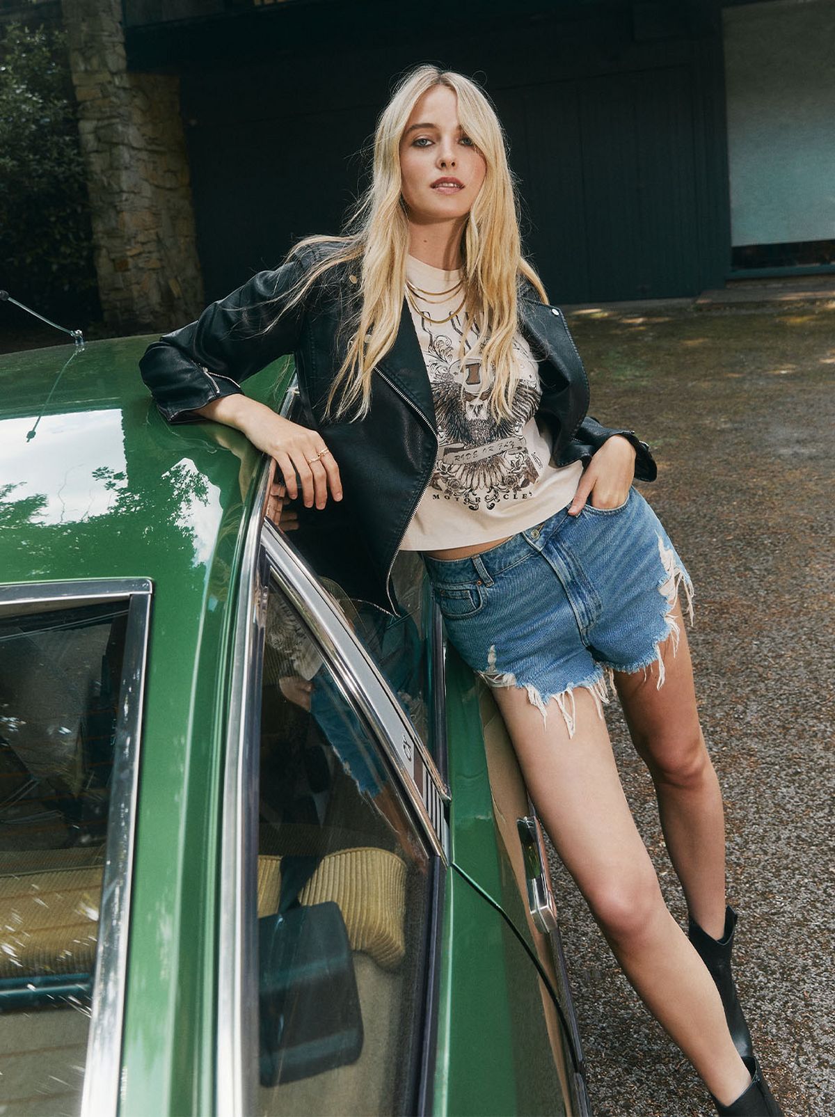 A women leans on a green classic car wearing a pair of sunglasses, a pair of black leather ankle boots, a black leather jacket and a pair of denim shorts