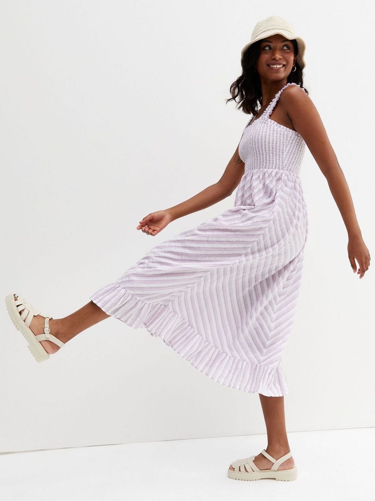 We see a women wearing a white and lilac patterned midi dress, facing to the side with her leg stretched forward as if stepping forwards, looking behind her and smiling