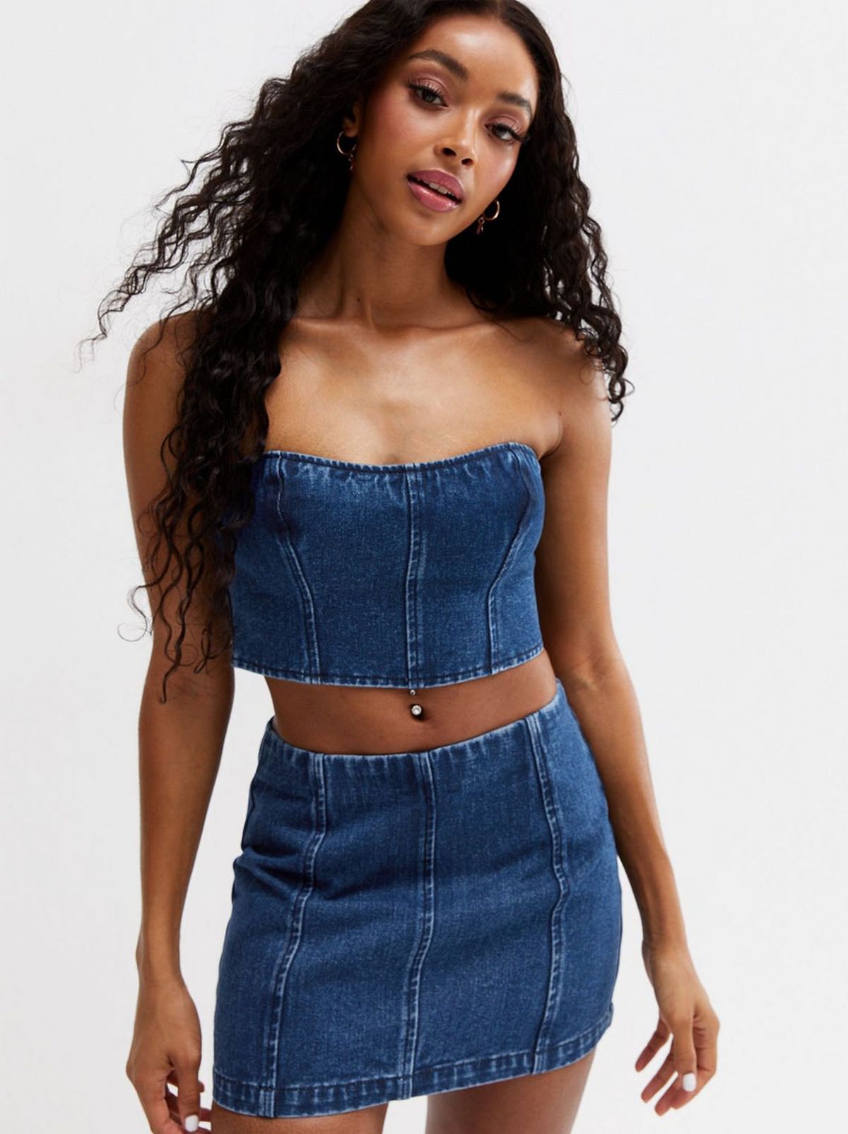 A women wears a blue washed denim mini skirt and bandeau top standing in front of a white background