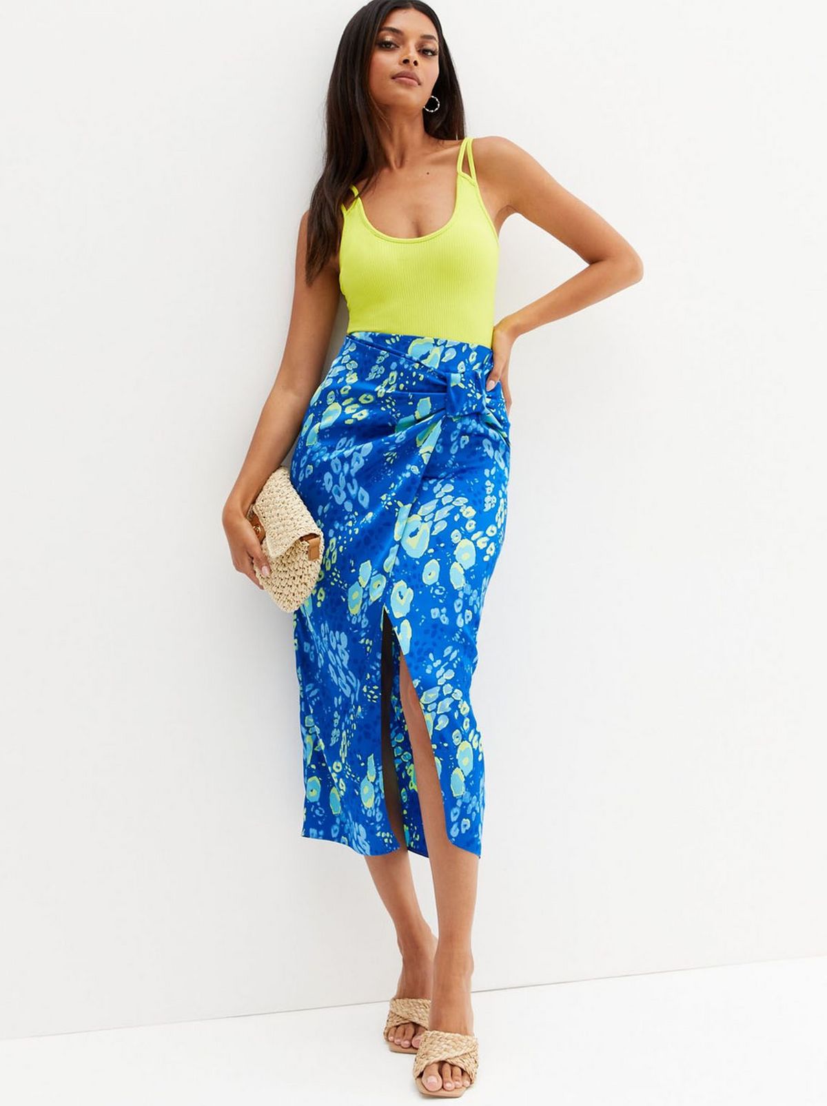 A women wears a blue patterned midi skirt and a lime green strappy top leaning on a white wall looking at the camera