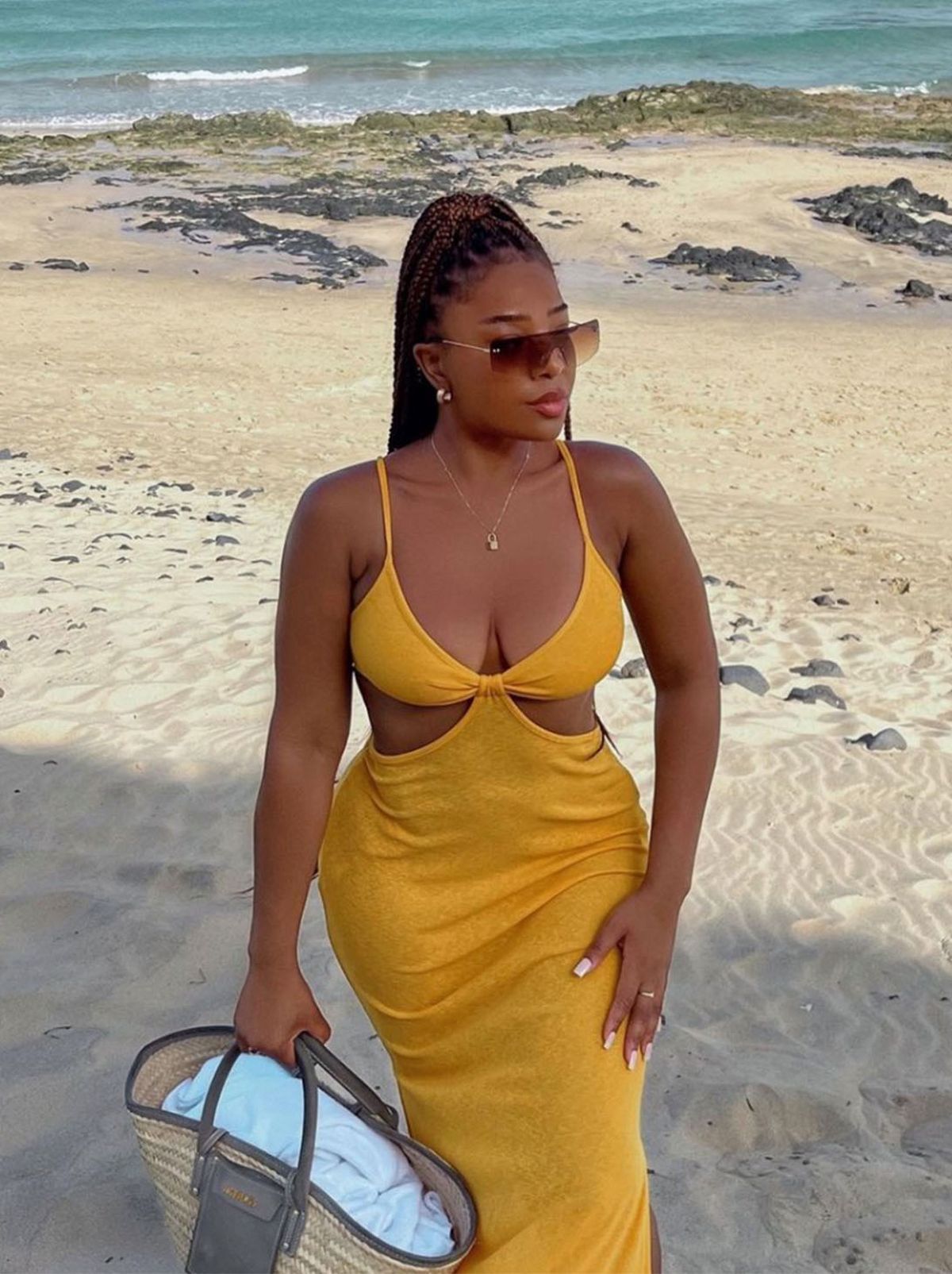 Sarah Holder stands on the beach wearing a yellow summer midi dress, holding a beach bag and looking into the distance