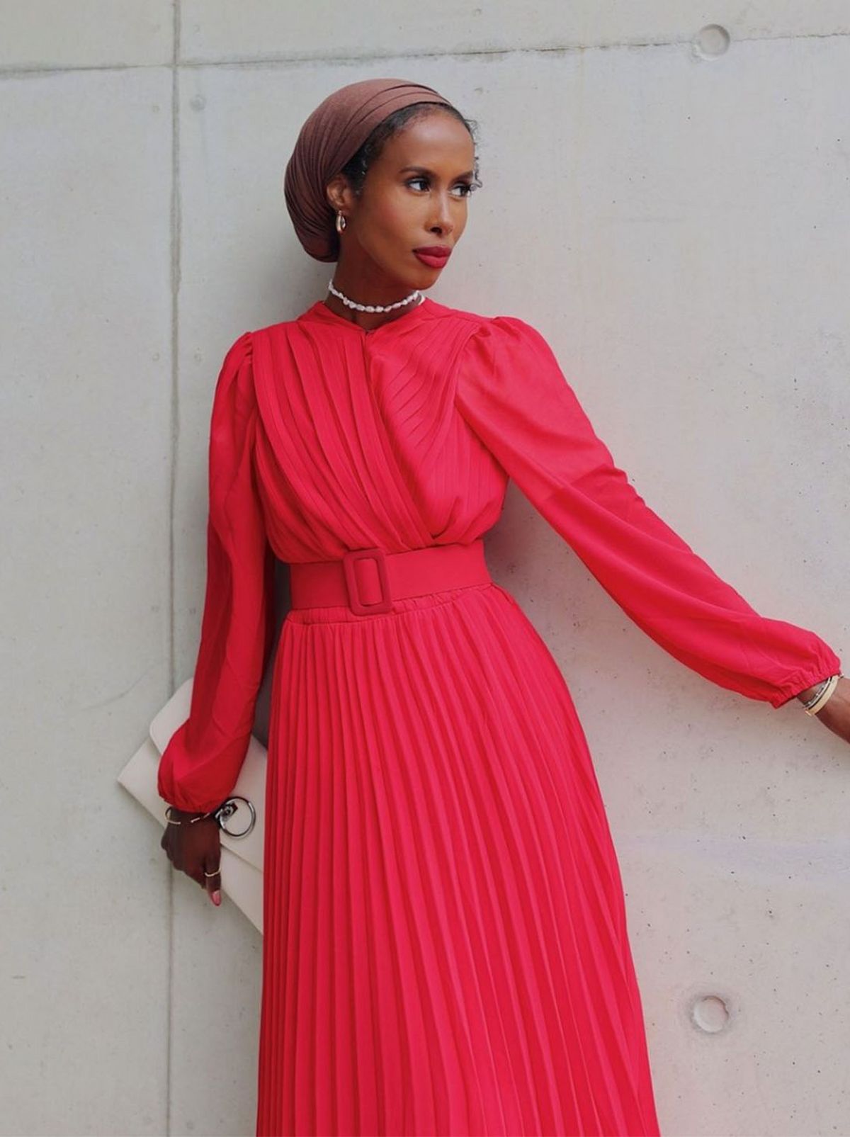 Basma Kahie stands leaning on a white wall looking into the distance, wearing a red midi dress and holding a white clutch