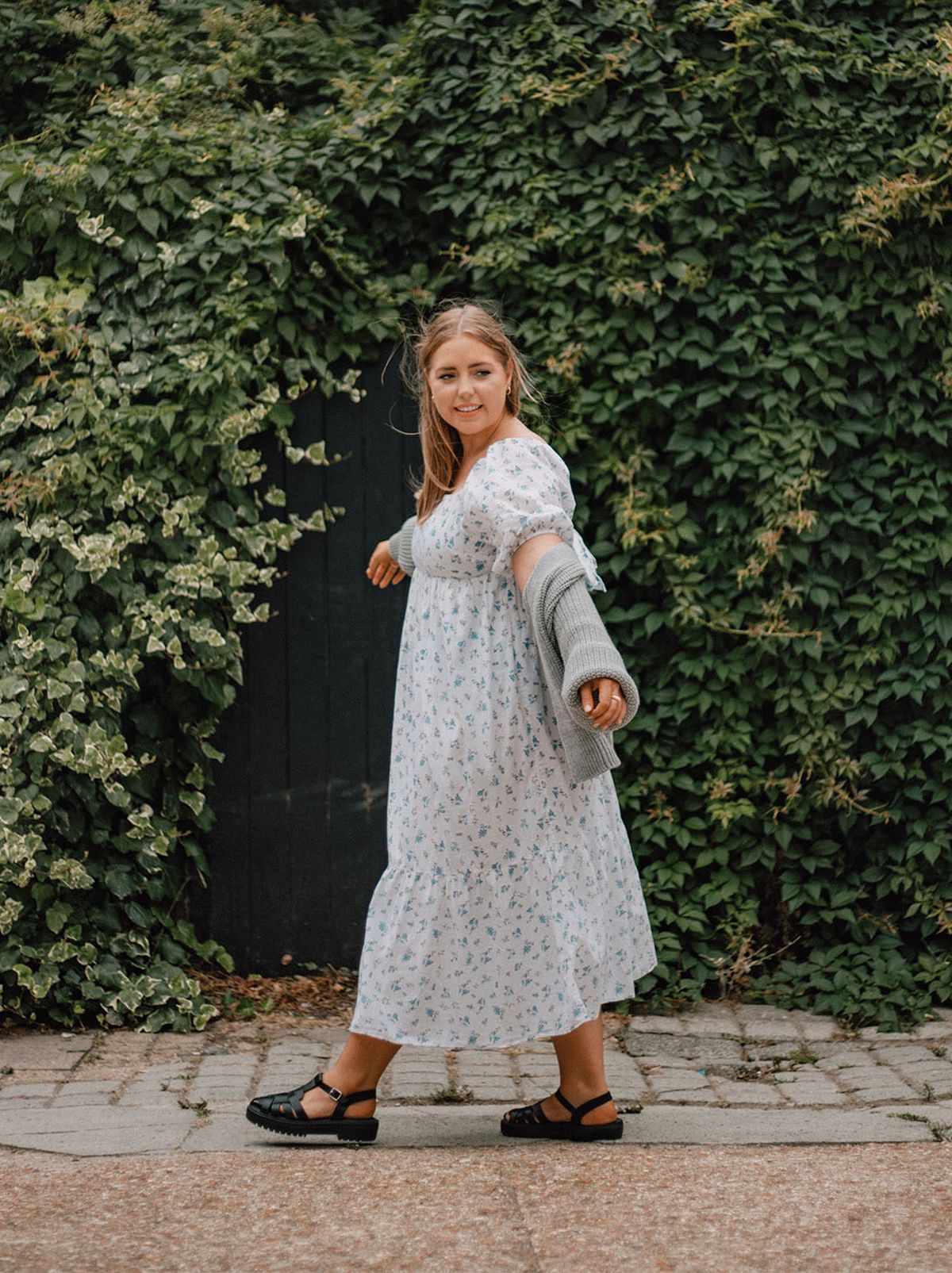 Poppy Deyes wears a white patterned maxi dress, holding her arms out to the side while walking down a street in front of a big hedge