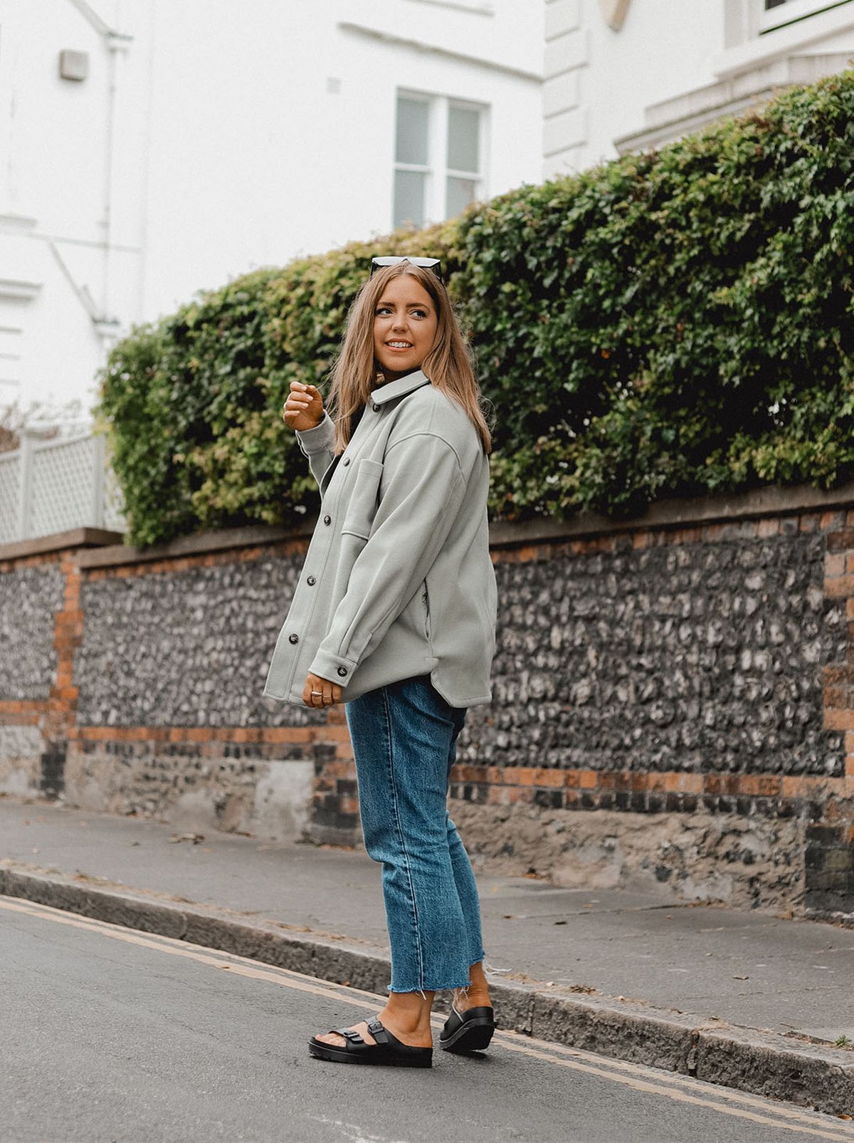 Poppy Deyes walks down a street, looking behind her and away from the camera while wearing a grey jacket and a pair of straight leg jeans