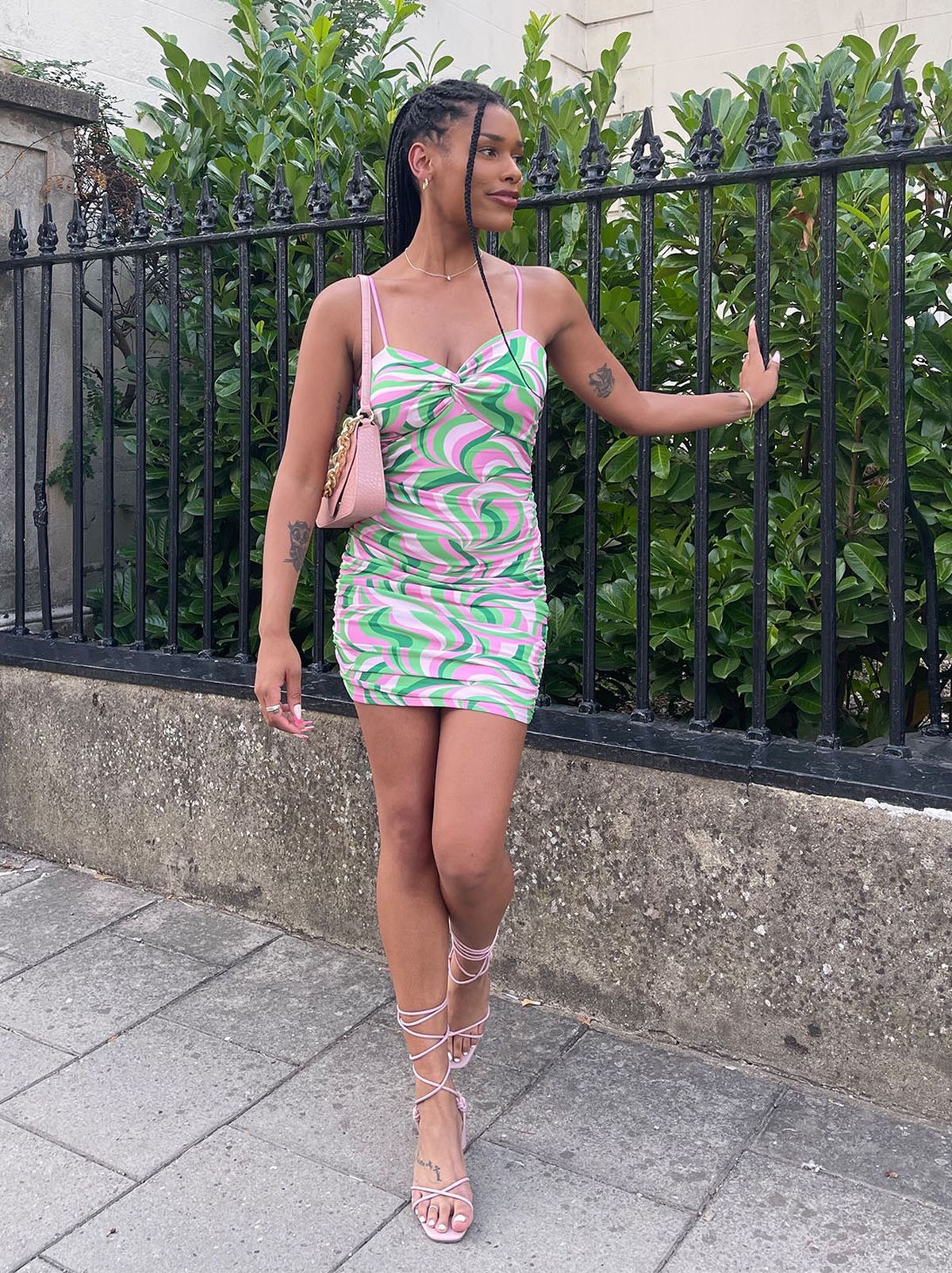 New Look's Jasmine stands on the pavement leaning on a railing wearing a pink and green bodycon mini dress from QUIZ