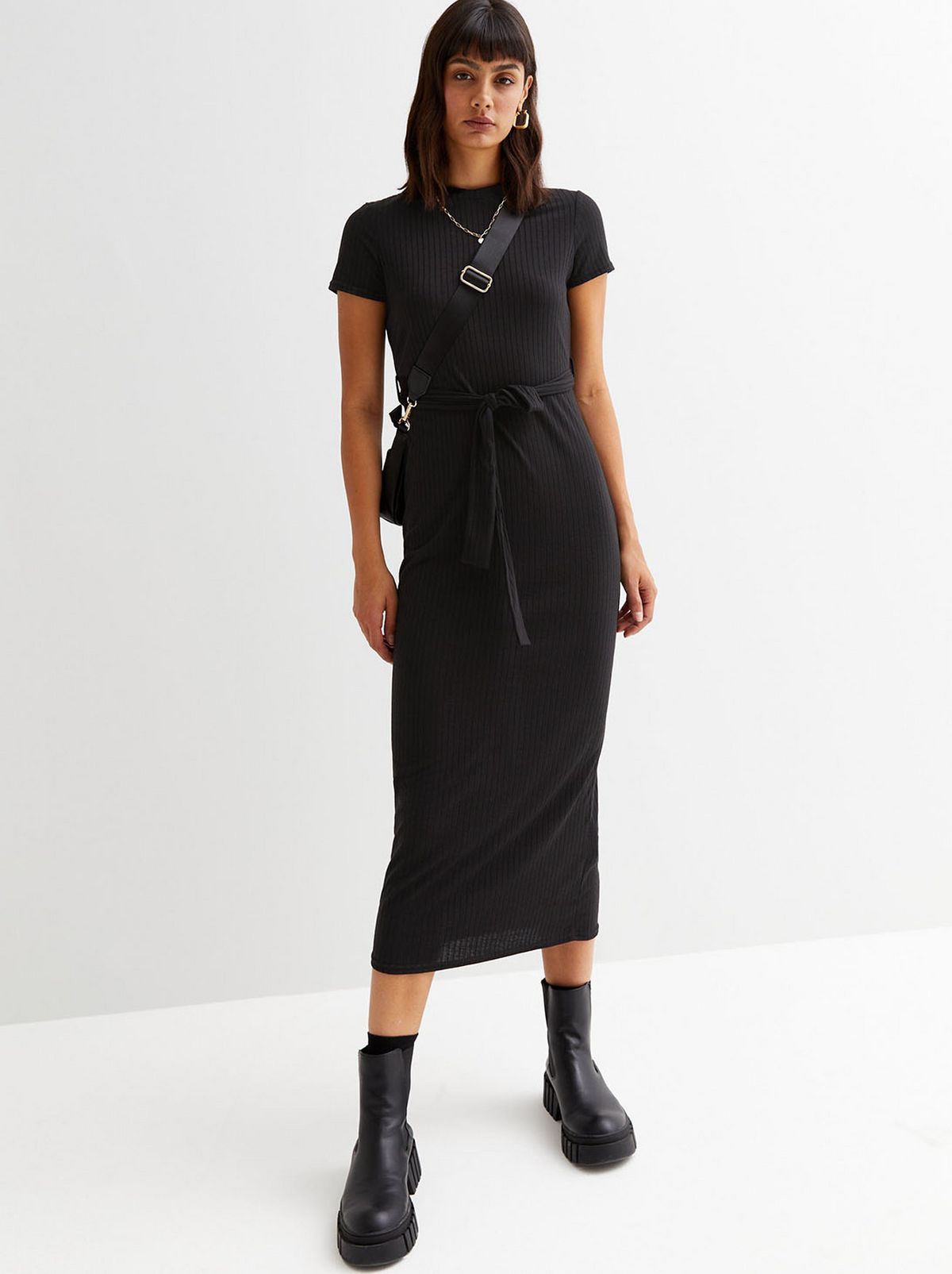 A model stands looking at the camera wearing a black ribbed midi dress and black ankle boots