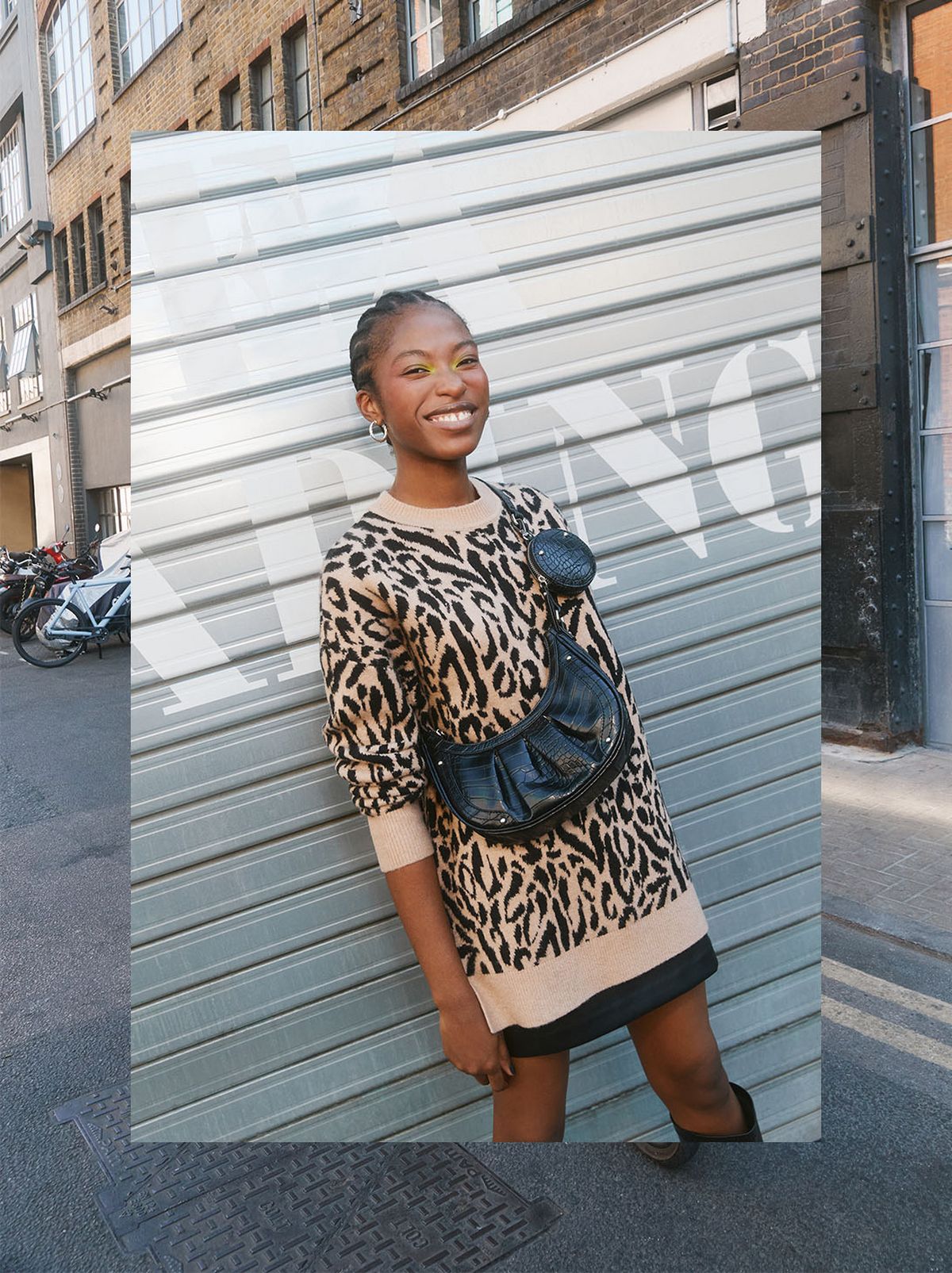 A model smiles at the camera standing in front of a metal wall, wearing a leopard print jumper, a black mini skirt and a black bum bag