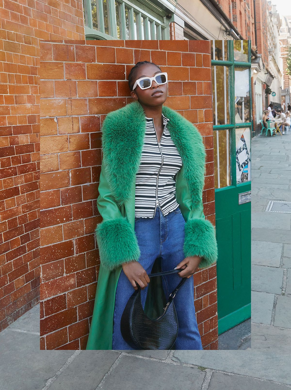 A model leans on a brick wall, wearing a pair of oversized white sunglasses, a green fluffy faux fur coat and a pair of blue jeans, holding a black handbag