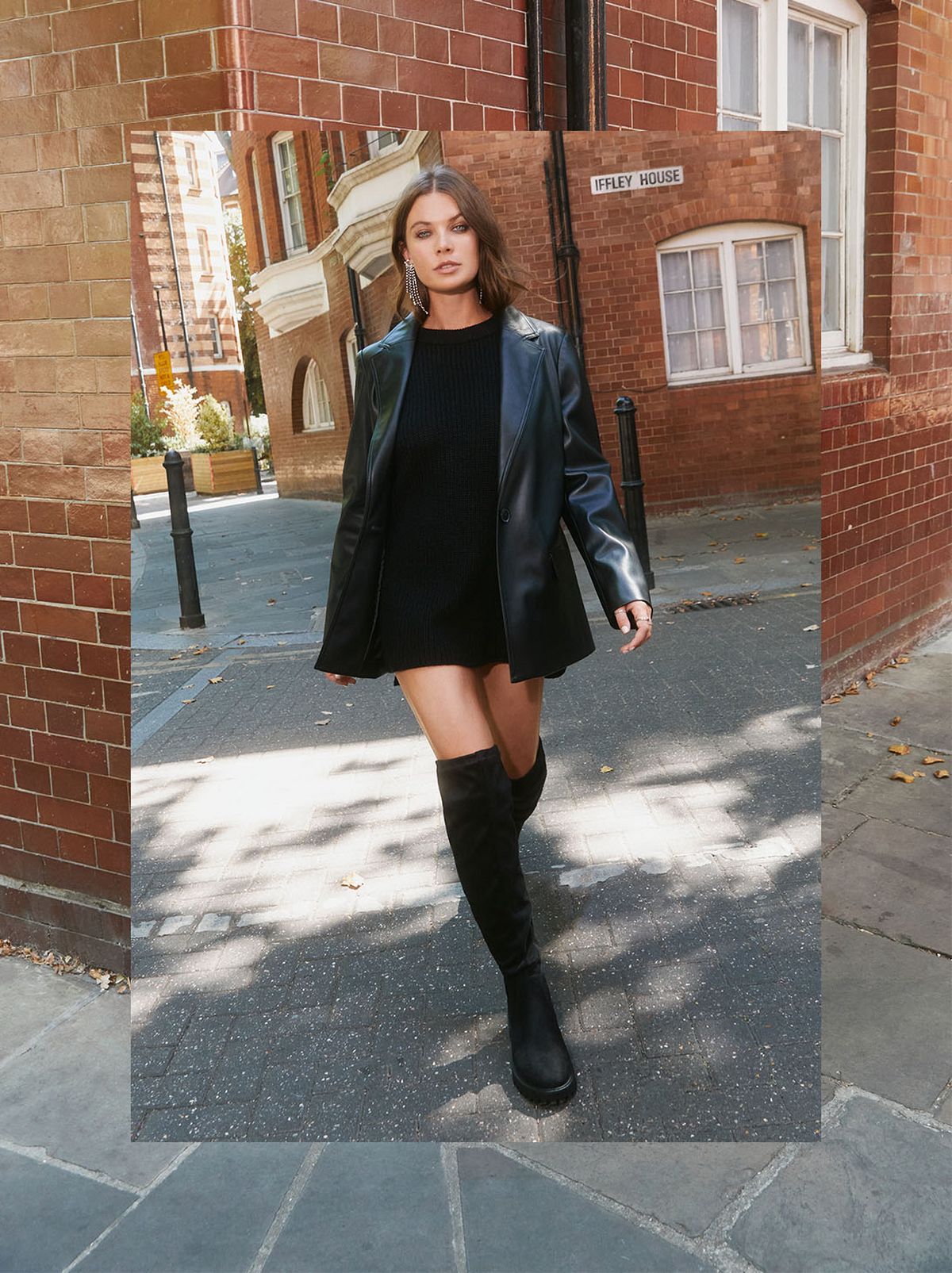 A model walks down a street wearing a black faux leather jacket, a black mini dress and a pair of knee high boots