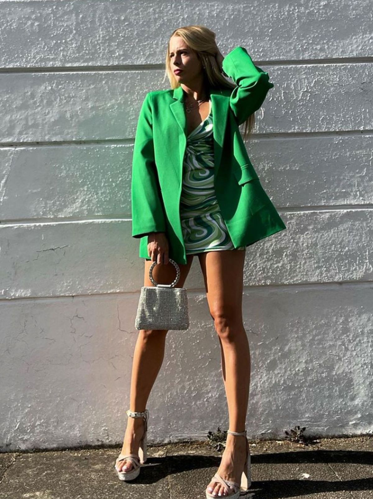 Emilie stands in front of a white wall, wearing a green blazer and a marble pattern mini dress, holding a handbag and wearing a pair of white heels