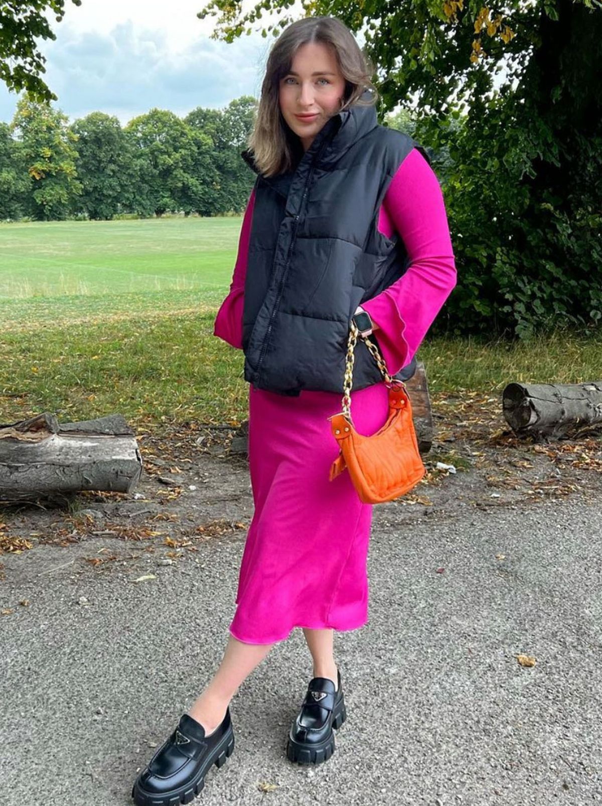 Charlotte stands with her legs crossed in front of some trees and a field, looking at the camera while wearing a hot pink long sleeve bodycon midi dress and a black gilet, holding a small orange handbag