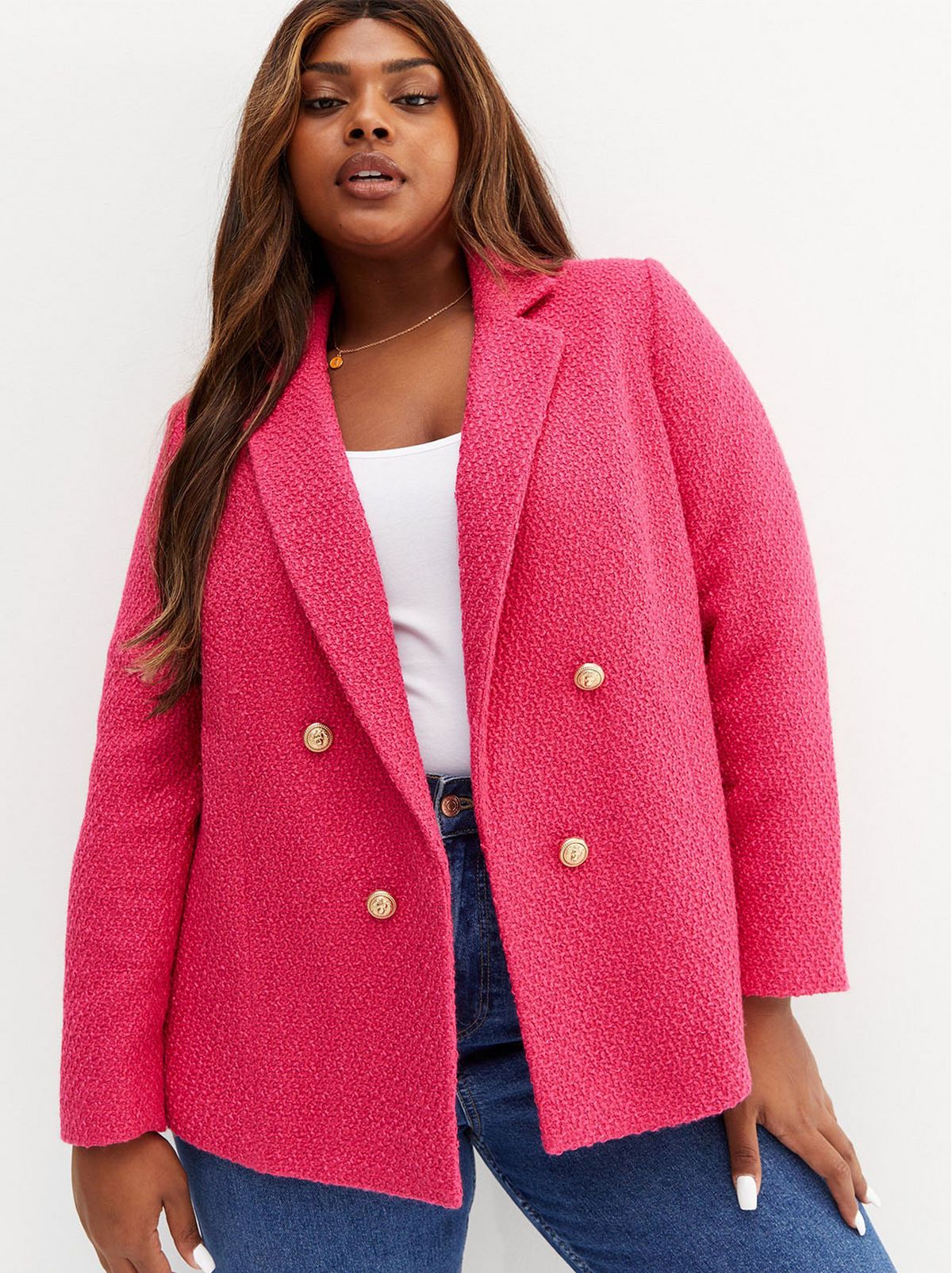 A model stands resting one hand on her leg, wearing a pink textured blazer, a white t shirt and a pair of acid washed blue jeans
