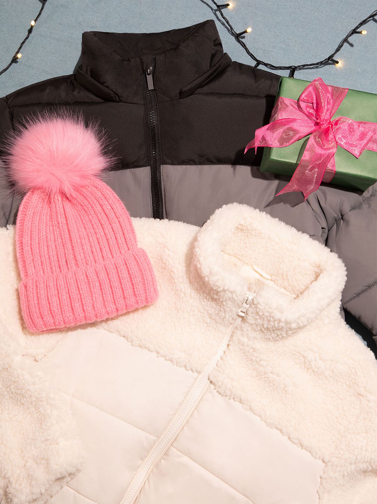 There is a cream fluffy jacket on top and a waterproof jacket under it, with a pink fluffy hat resting on top, in the top right corner is a green present with a pink bow, and some lights in the top right corner