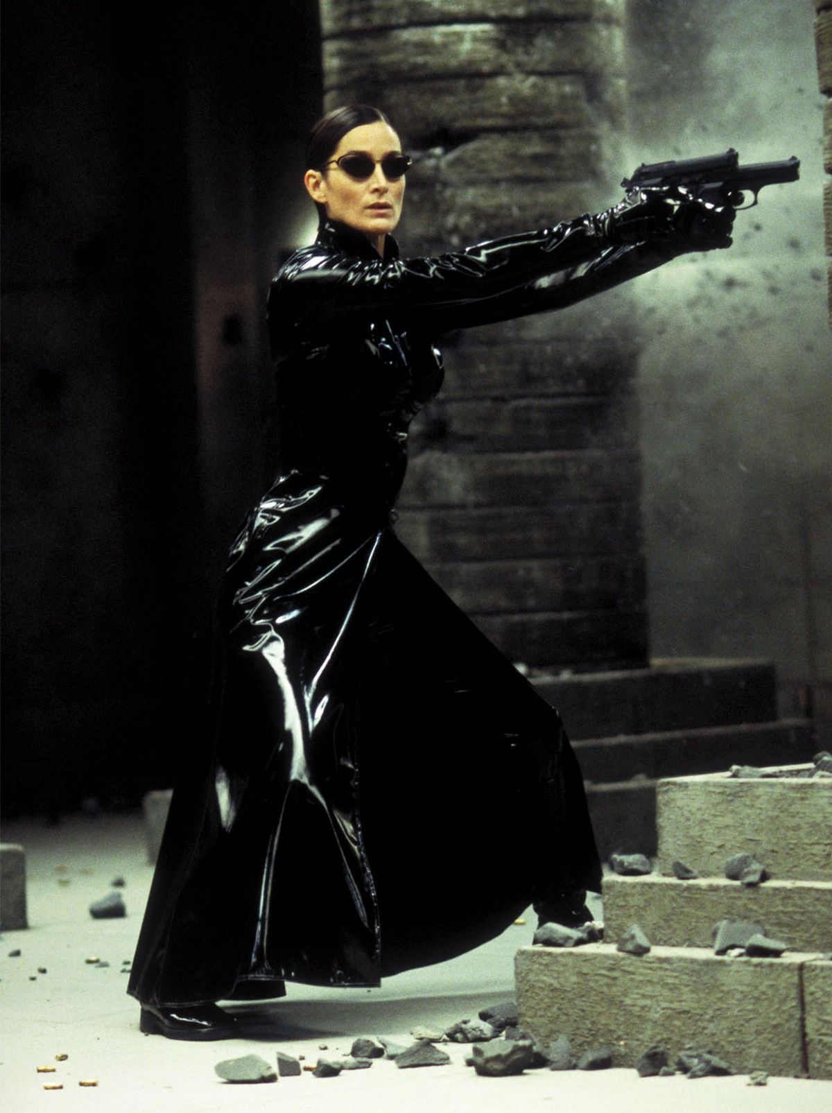 Trinity stands in a corridor, shooting at something out of shot, while wearing a long black jacket, black leather look trousers and a pair of black sunglasses