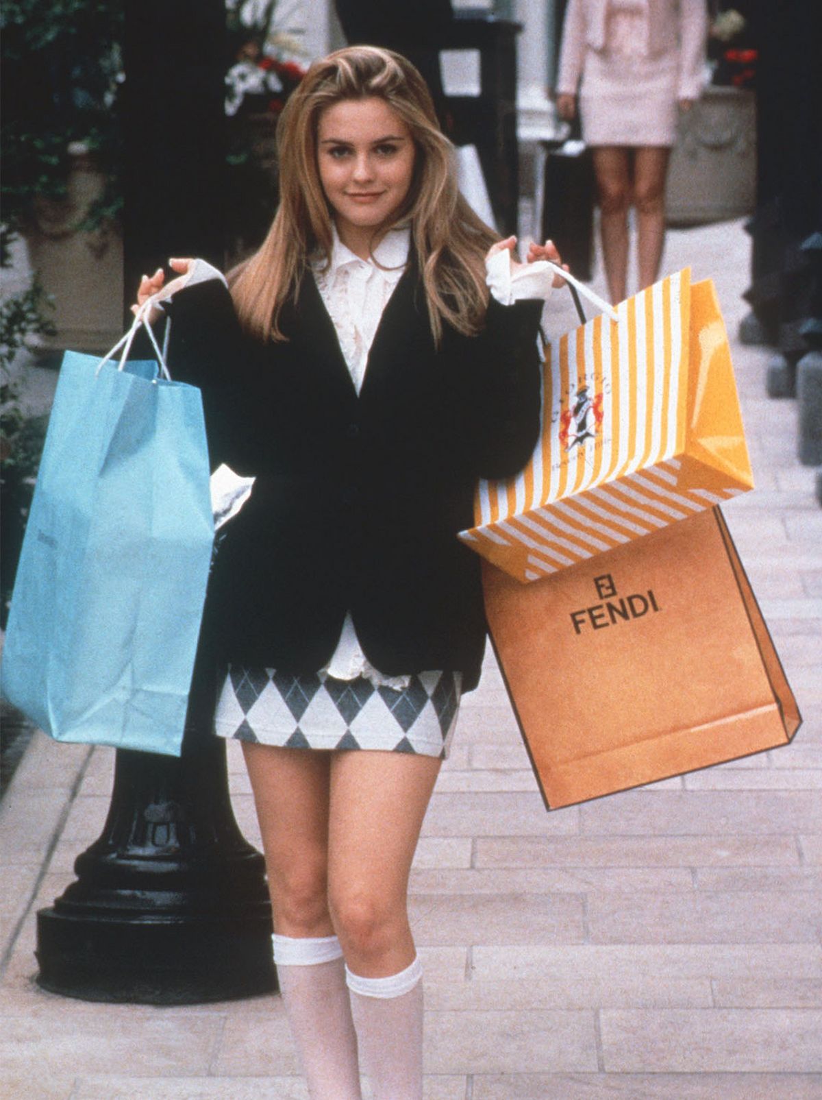 Cher Horowitz stands on a pavement with several bags thrown over either shoulder, smiling at the camera while wearing a black cardigan, a collared shirt and a pleated skirt