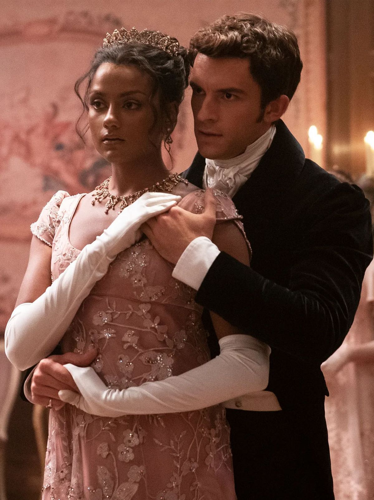 Kate Sharma looks ahead of her with a man standing behind her holding her hand, wearing a pair of long white gloves and a pink patterned dress