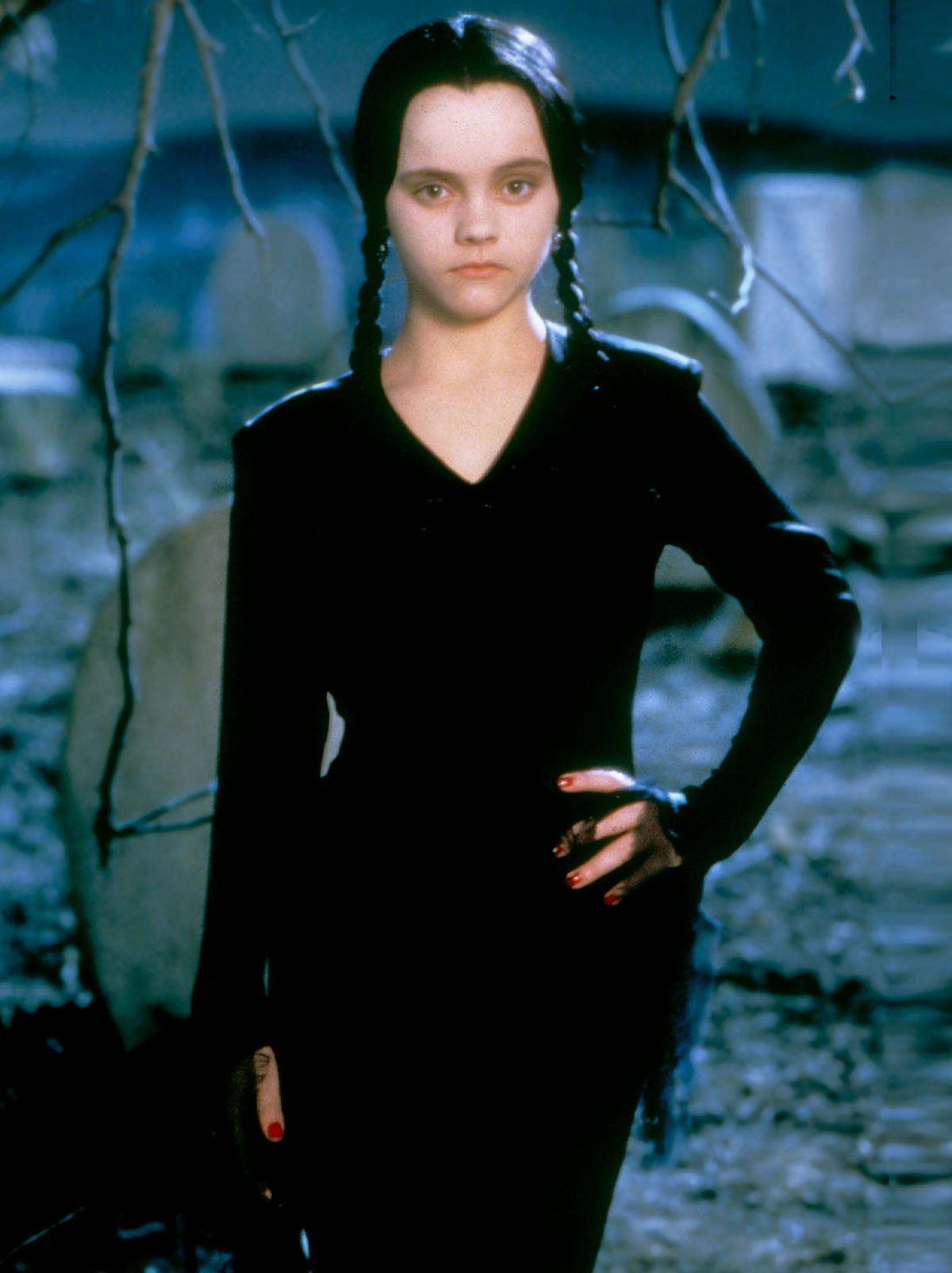 Wednesday Addams stands in a graveyard with a hand on her hip, wearing a black long sleeve dress 