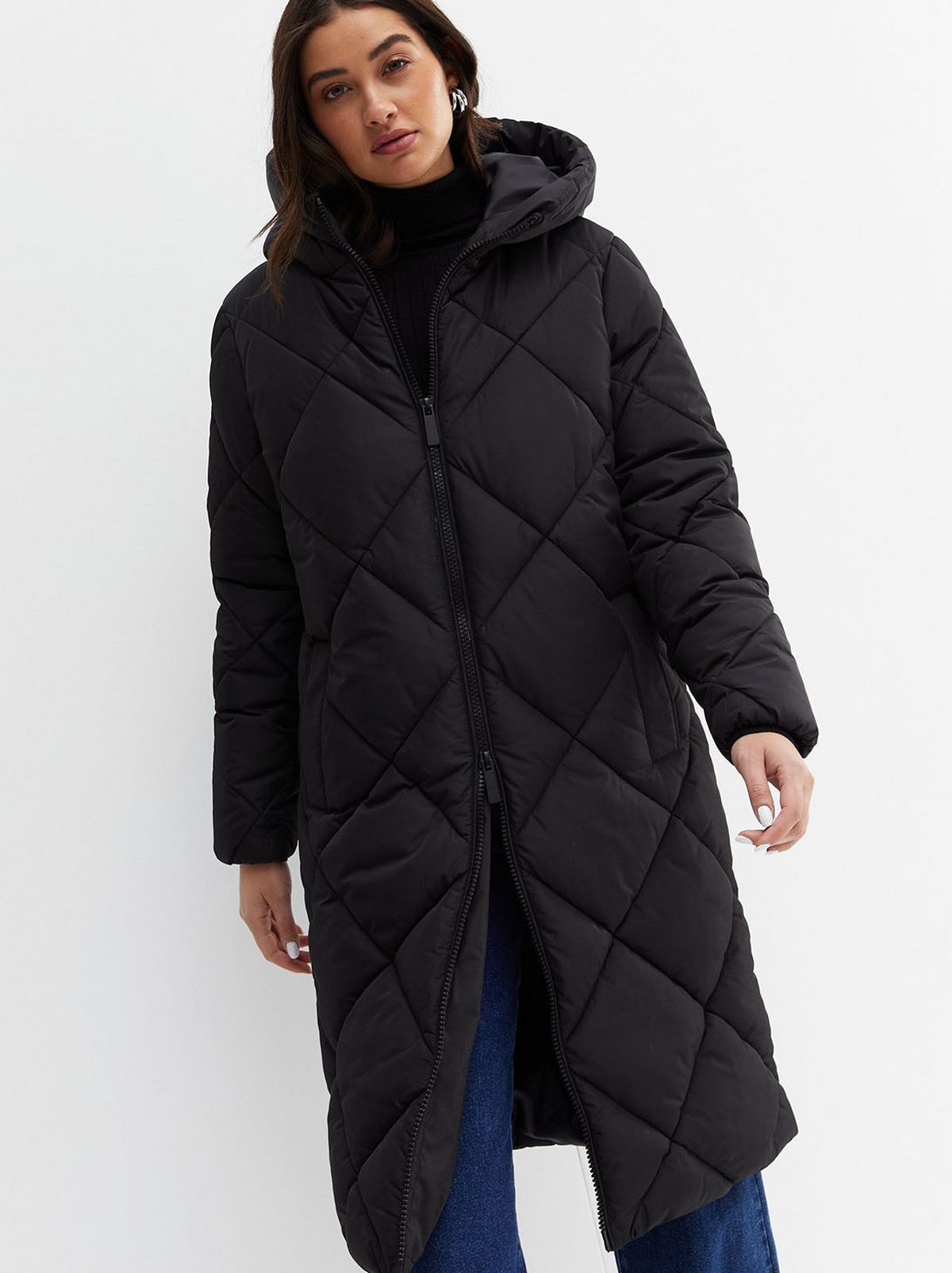 A model stands looking to the side at the floor in front of a blank background, wearing a pair of black trousers, a white shirt and a black chunky knit crew neck vest