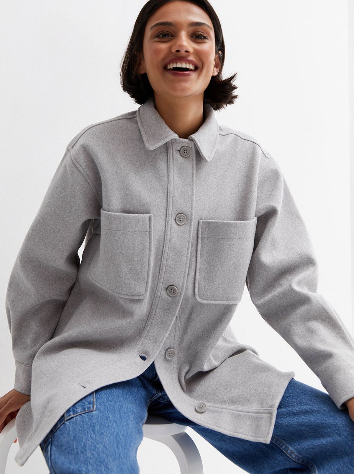 A model sits on a stool, smiling at the camera while wearing a pair of acid washed denim jeans and a pale grey double pocket shacket in front of a blank background
