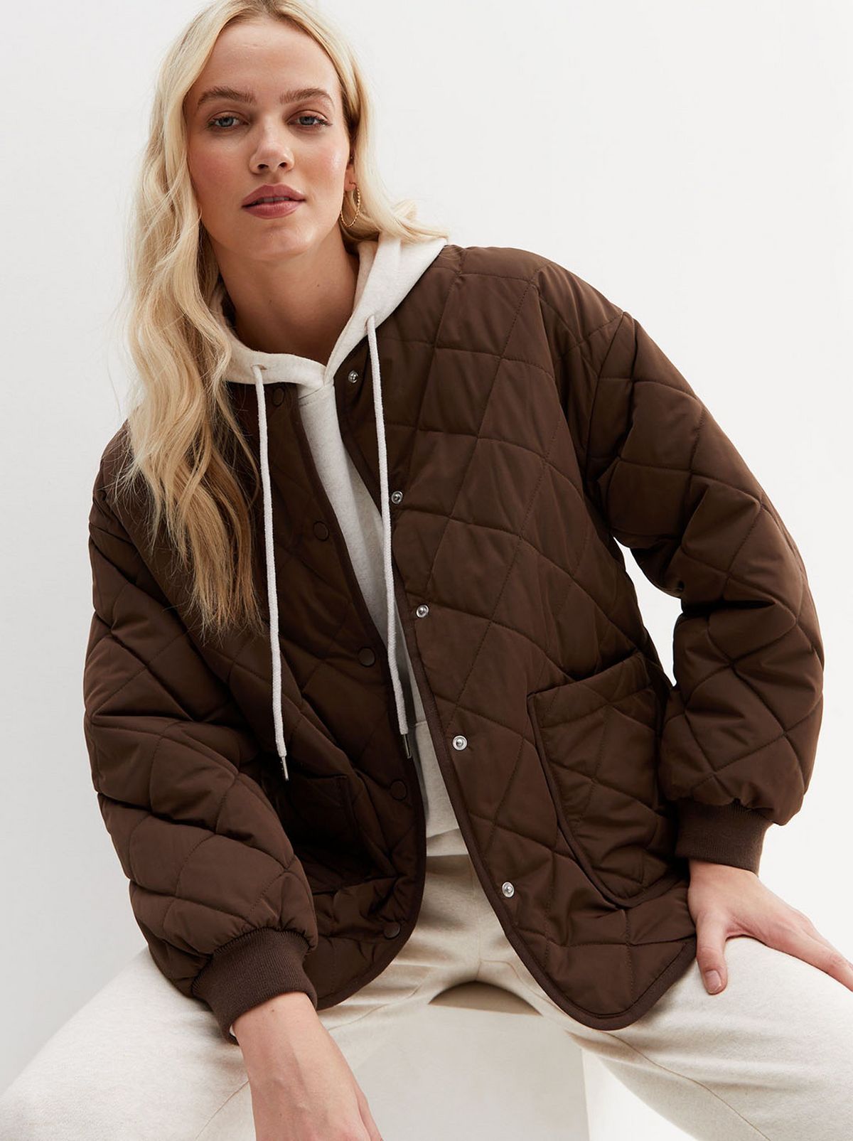 A model sits on a stool resting their arm on their thigh, looking at the camera in front of a white background wearing a dark grown quilted jacket, a pair of cream trousers and cream jumper