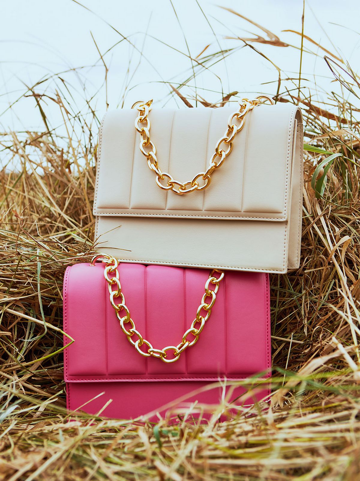 Two faux leather bags are stacked on top of each other in a field of straw, one bright pink and one is white, both with gold chains