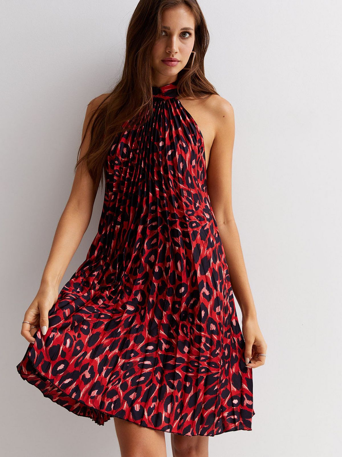 A model looks at the camera in front of a white background, holding the hem of their red leopard print pleated mini halter dress