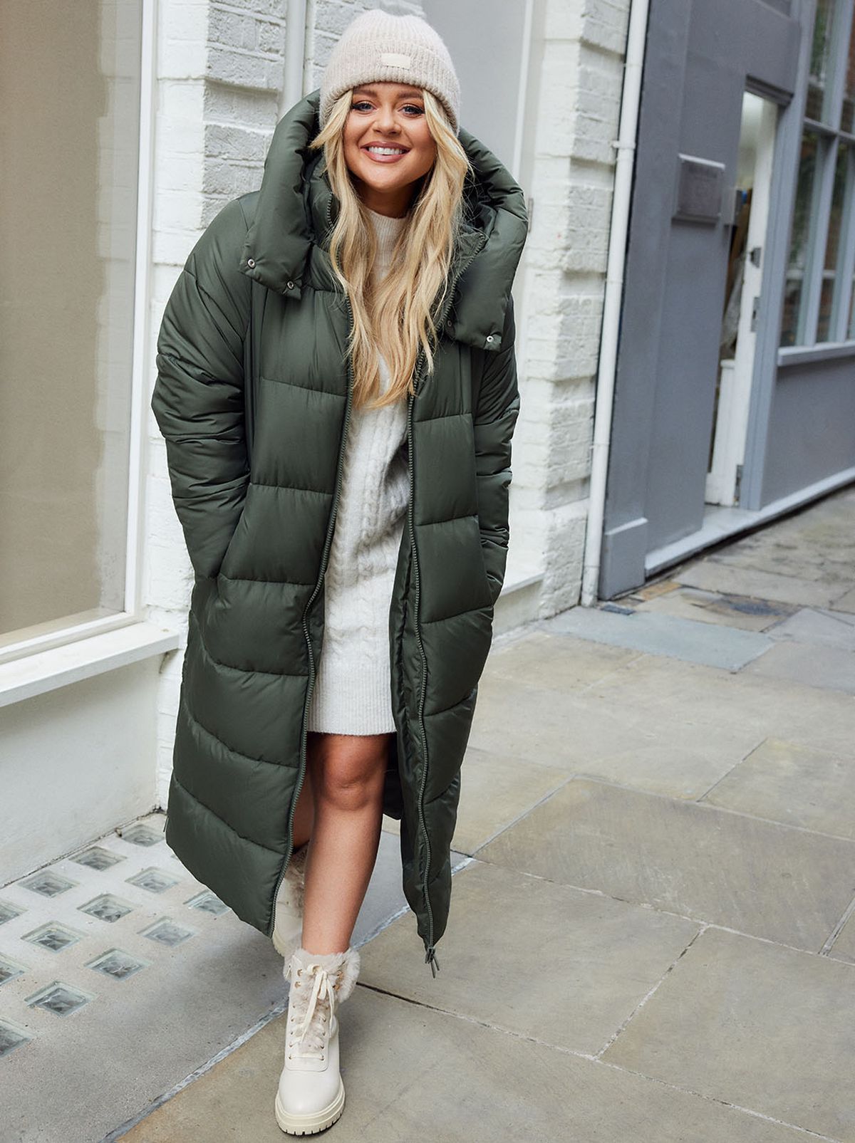 Emily Atack walks towards the camera while smiling at the camera on a city street, wearing a cream knitted dress and olive longline puffer coat
