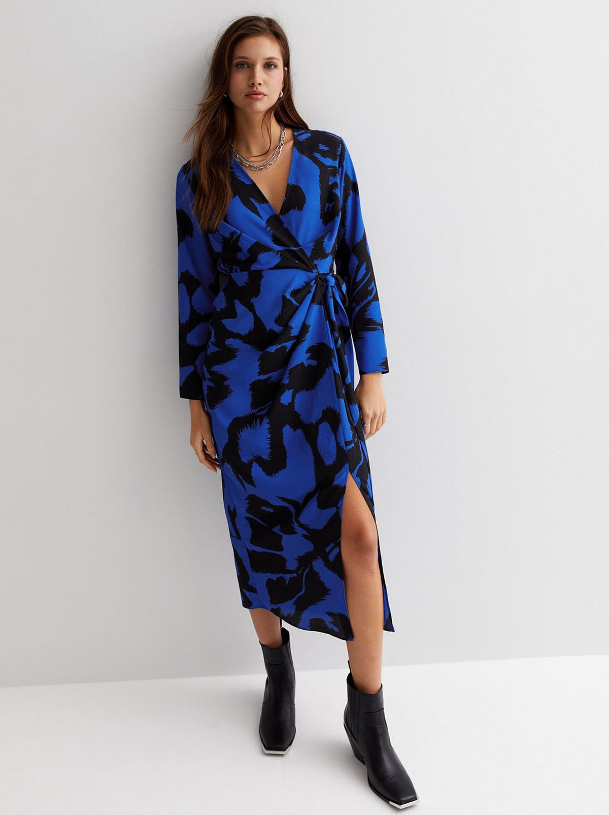 A model is standing looking at the camera in front of a white background wearing a blue patterned wrap midi dress and a pair of black boots