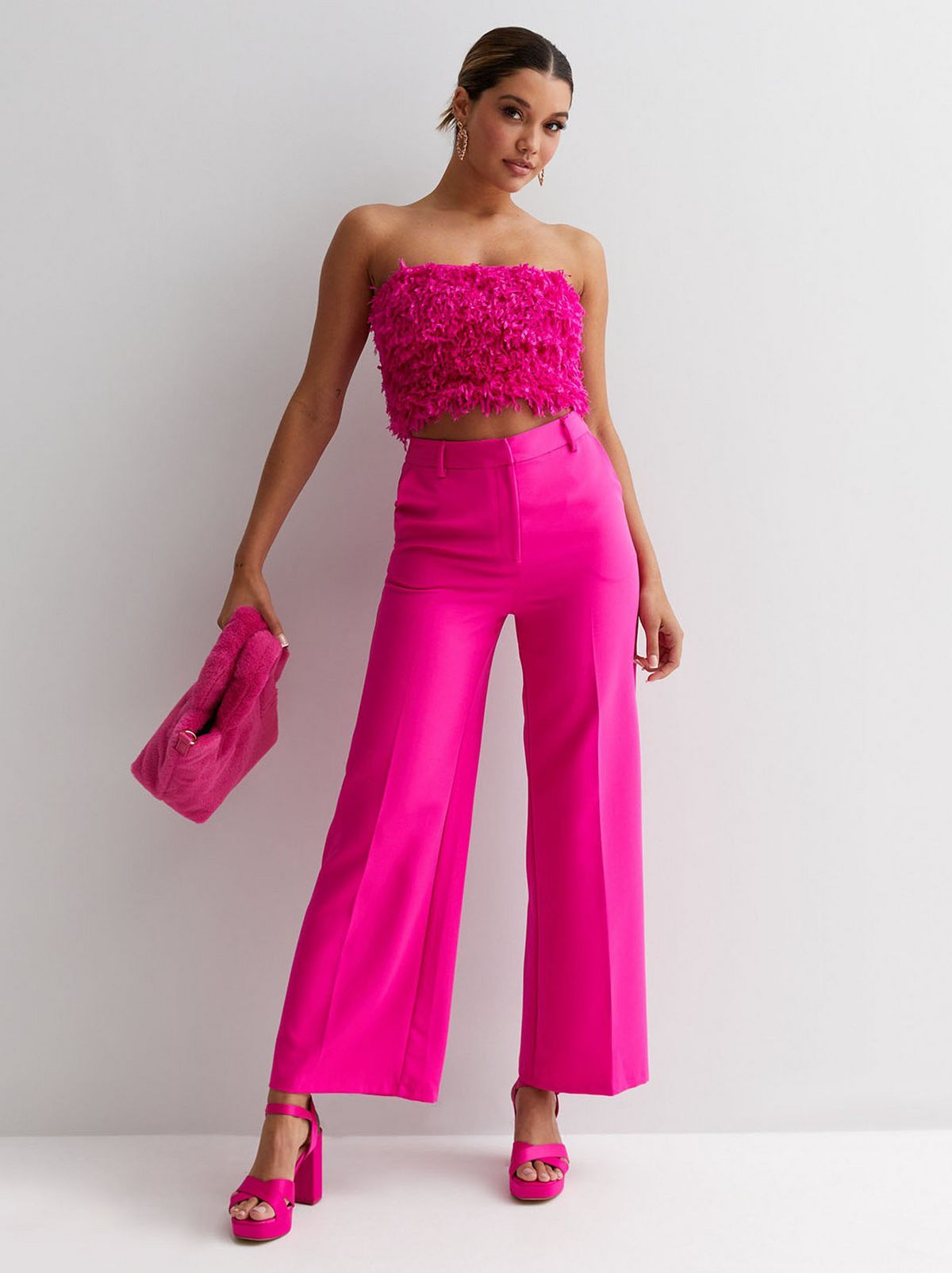 A model stands in front of a blank background looking at the camera wearing a bright pink faux feather bandeau and a pair of hot pink trousers
