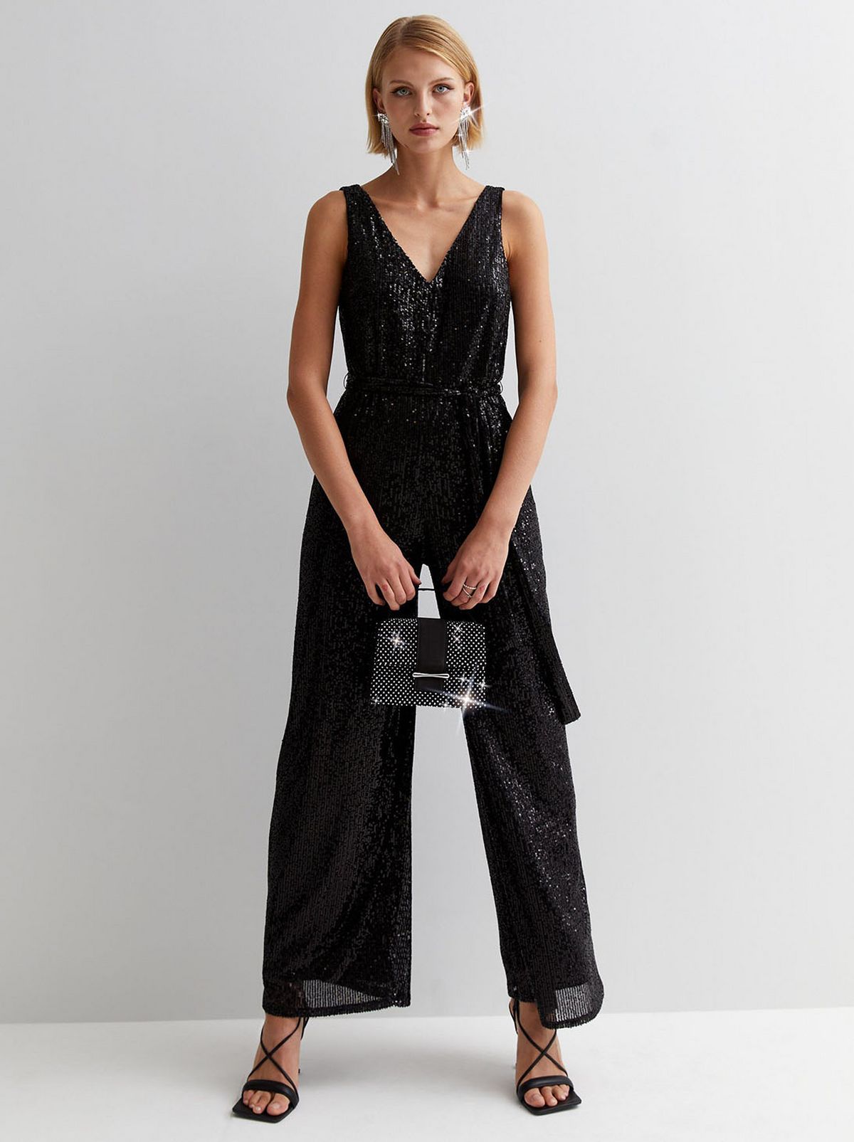 A model stands in front of a blank background wearing a black sequin sleeveless jumpsuit