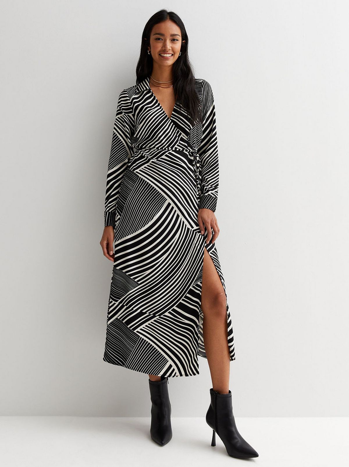 A model stands in front of a blank background smiling at the camera while wearing a black patterned long sleeve midi dress
