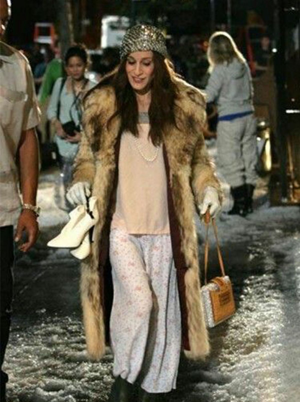 A women walks towards the camera outside on an icy floor wearing a long fur coat and a silver hat while holding a pair of cream shoes and a brown handbag