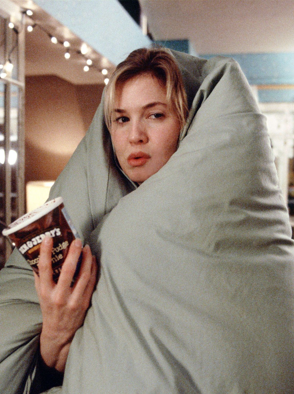 A women sits covered in a large duvet while holding a tub of ice cream with a spoon inside it, there is a patterned blue wallpaper on the left and a shelf just above it